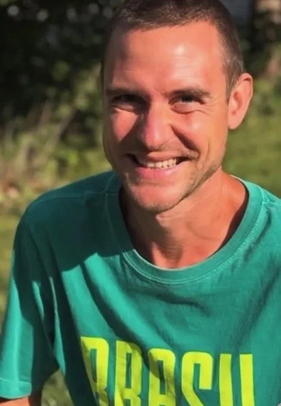 A smiling man wearing a green t-shirt with yellow text, outdoors on a sunny day, with trees in the background.