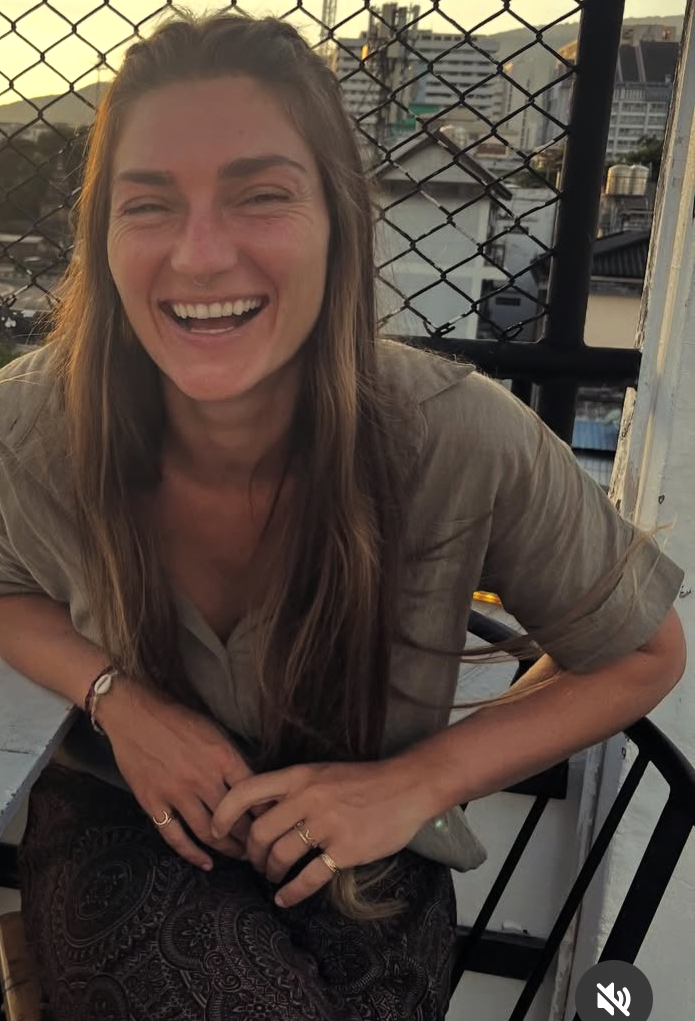 A woman with long brown hair and a big smile, sitting outdoors near a black metal railing with a cityscape in the background during sunset.