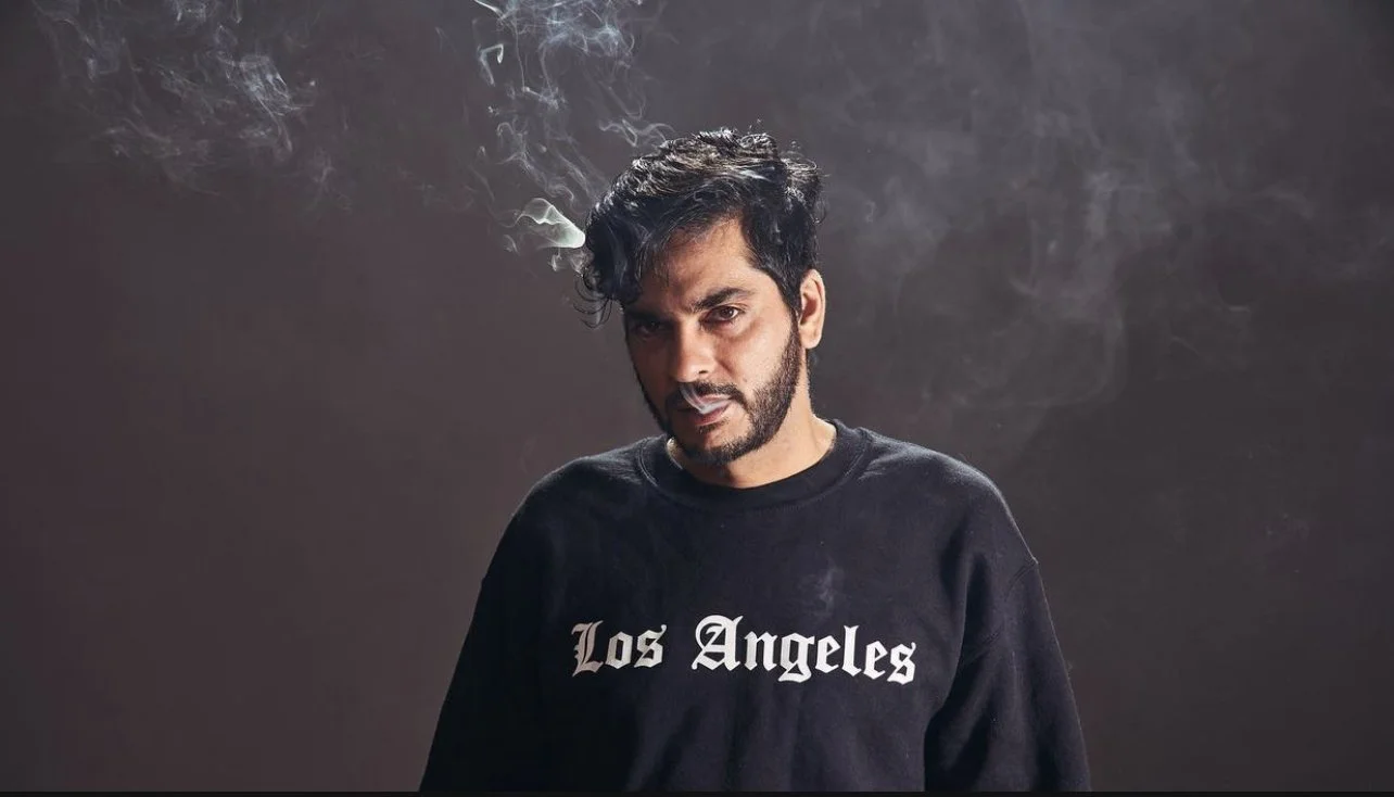 Man with beard wearing a "Los Angeles" sweatshirt, surrounded by smoke.