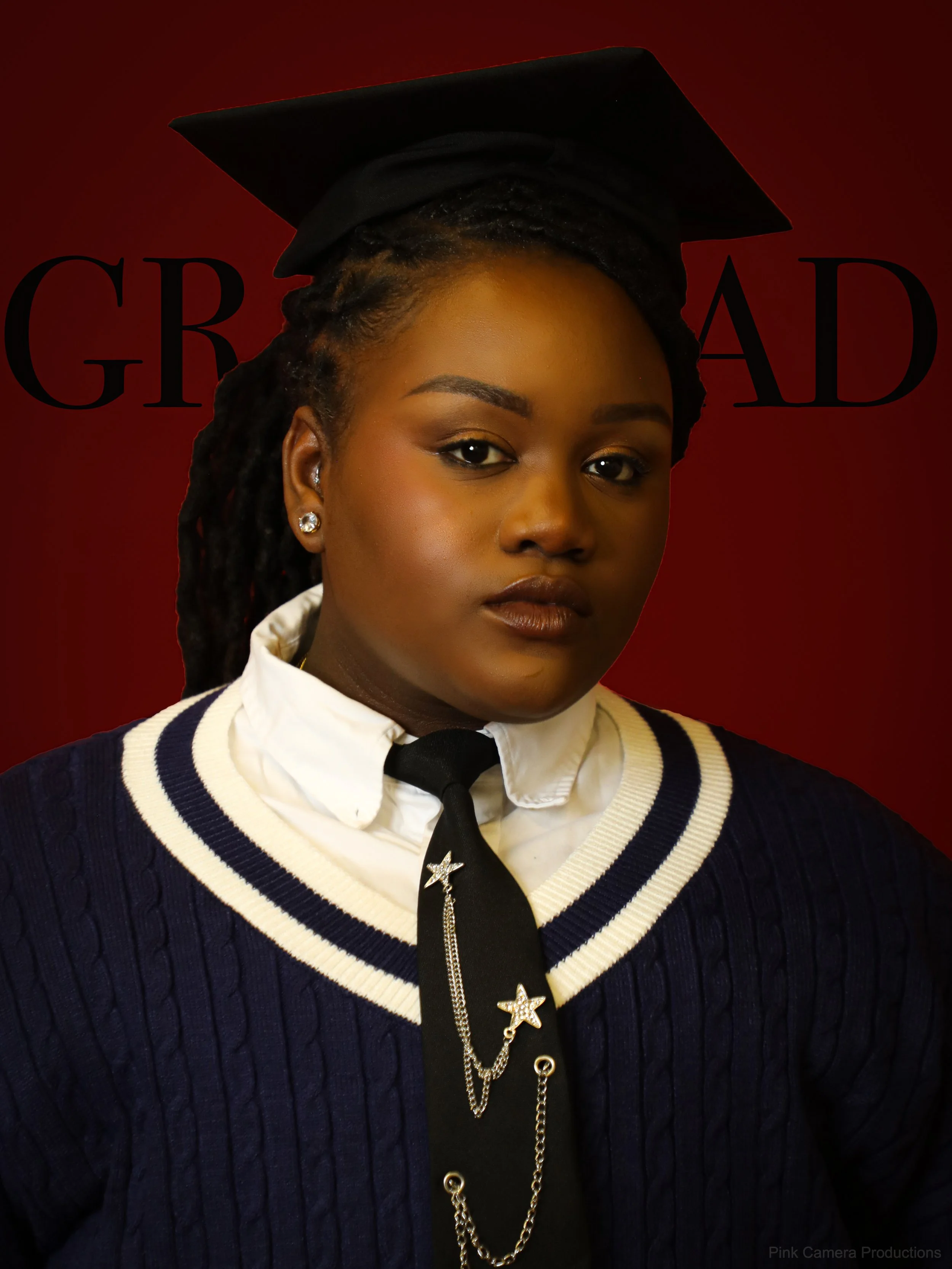 A young woman in graduation attire with a mortarboard, wearing a white shirt and a dark navy sweater with white trim, with a black tie decorated with star-shaped pins and chains. The background is dark red with partially visible large black text.