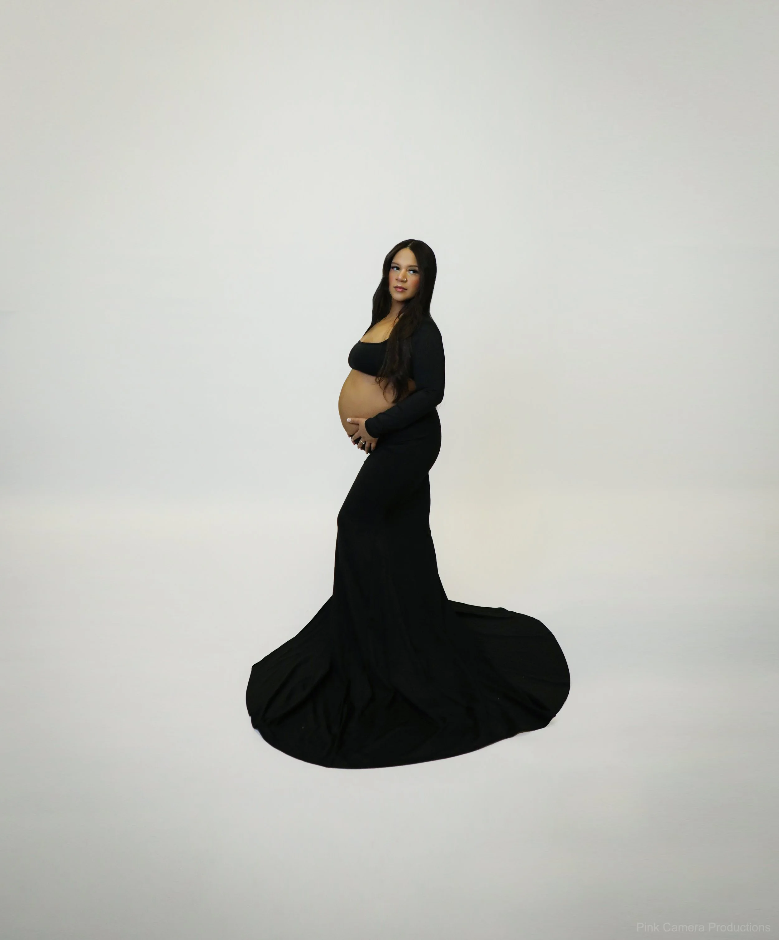 Pregnant woman in a black dress holding her belly, standing against a plain white background.