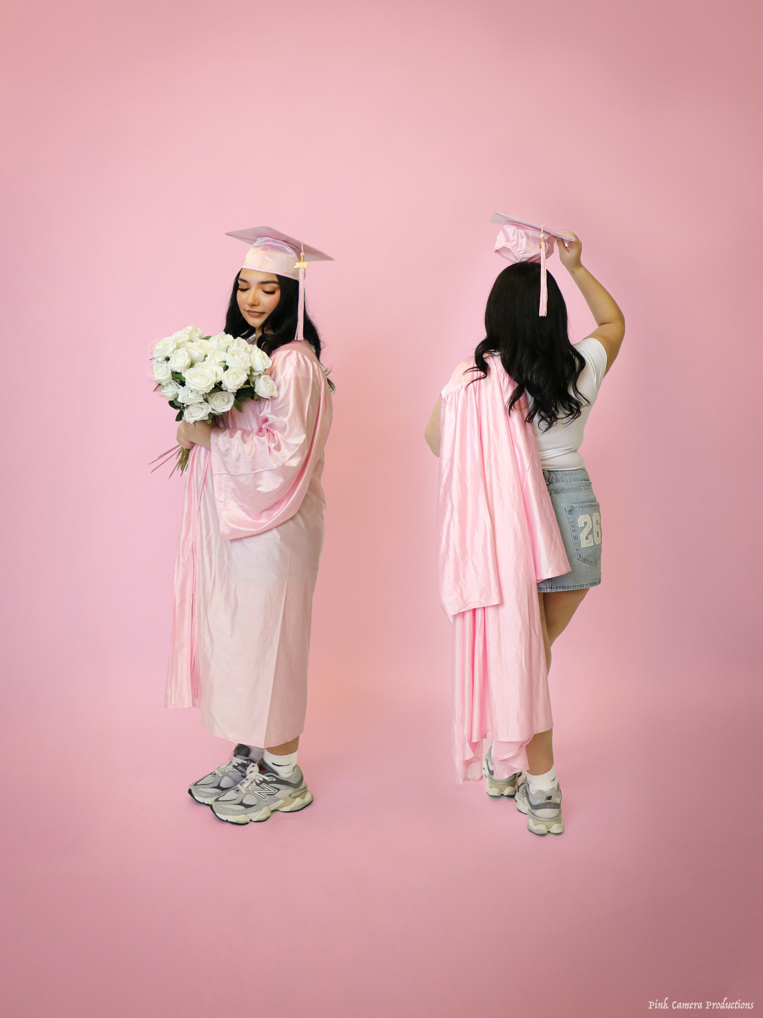 Two young women in pink graduation gowns and caps, one holding a bouquet of white roses. The woman on the left is facing slightly to the right, looking down at roses, and the woman on the right is touching her cap with her back to the camera. They ar