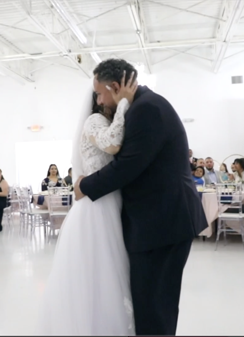 Couple dancing closely at a wedding reception, surrounded by seated guests in a bright, modern venue.