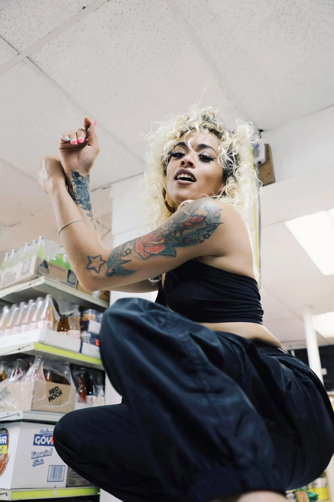 A woman with blonde curly hair, tattoos on her arm, and dark makeup, is squatting in a store aisle, looking at the camera, with shelves of bottles and grocery products in the background.