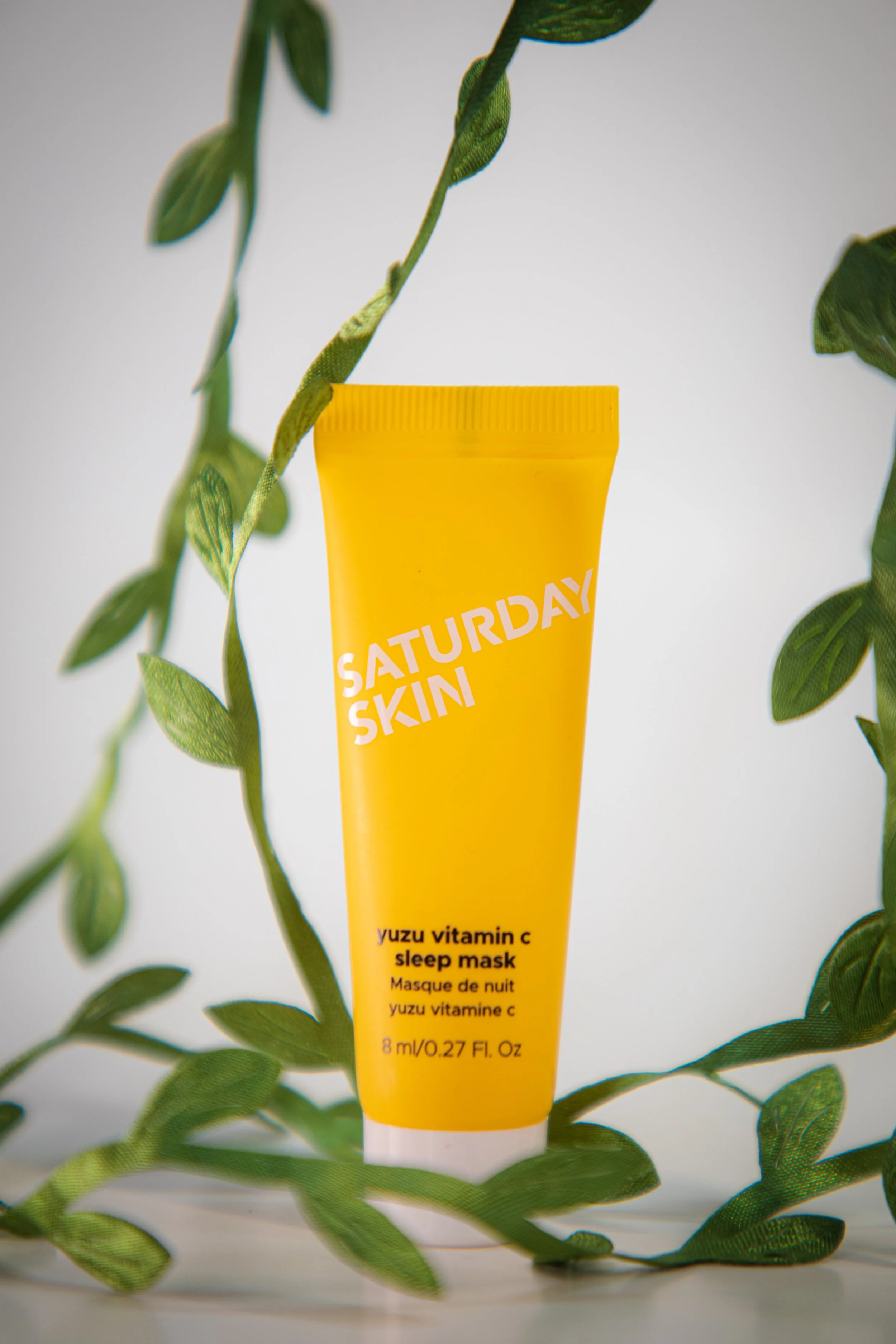 A yellow tube of Saturday Skin yuzu vitamin C sleep mask surrounded by green leaves on a light background.