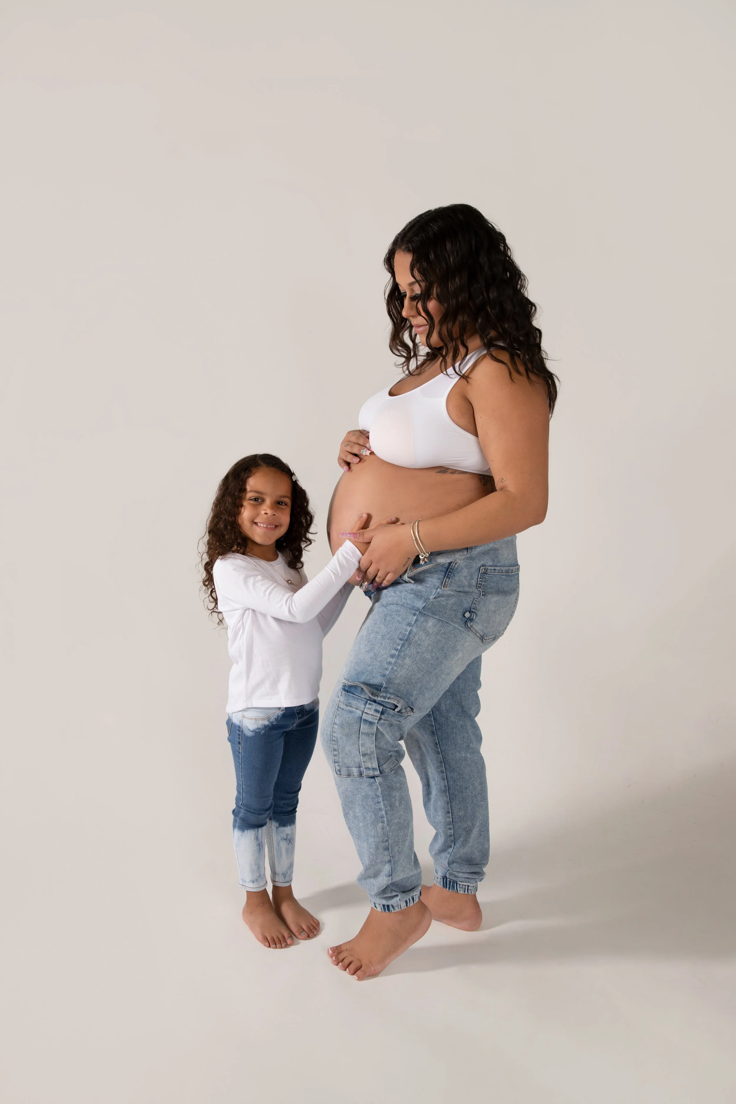 A pregnant woman in a white crop top and jeans standing next to a young girl in a white long sleeve shirt and jeans touching her pregnant belly and smiling.