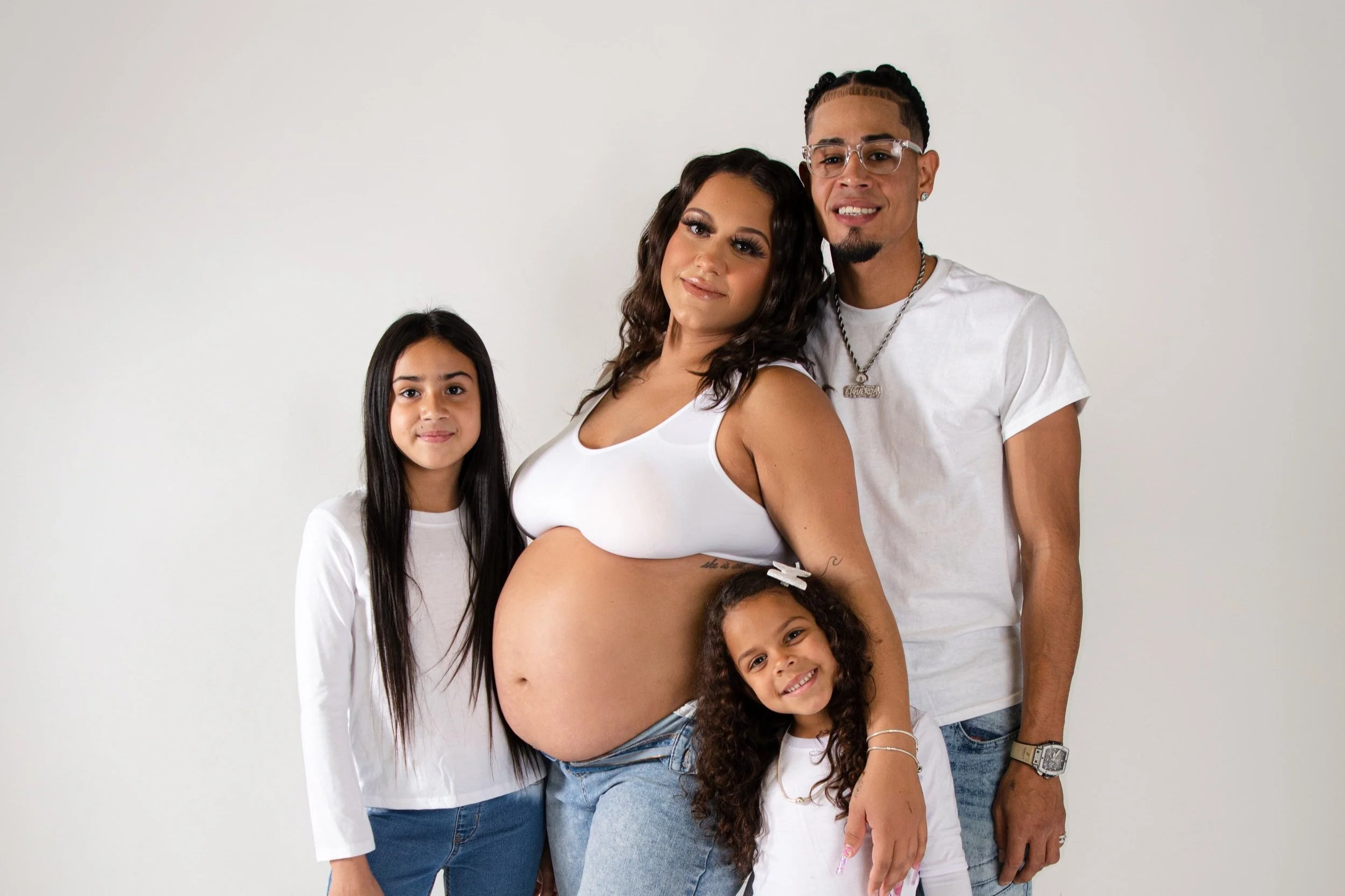 A pregnant woman with curly hair and a smile, standing with her family against a plain white background. She is wearing a white crop top and jeans, with her hand on her belly. To her left are two young girls, one with long black hair wearing a white 