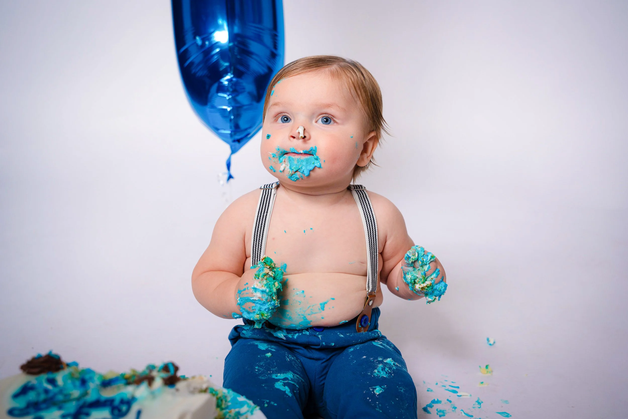 A young child with blue eyes and light brown hair, wearing blue pants and suspenders, sitting shirtless in front of a white background, covered in blue frosting, with a partially eaten birthday cake and a blue balloon floating behind him.
