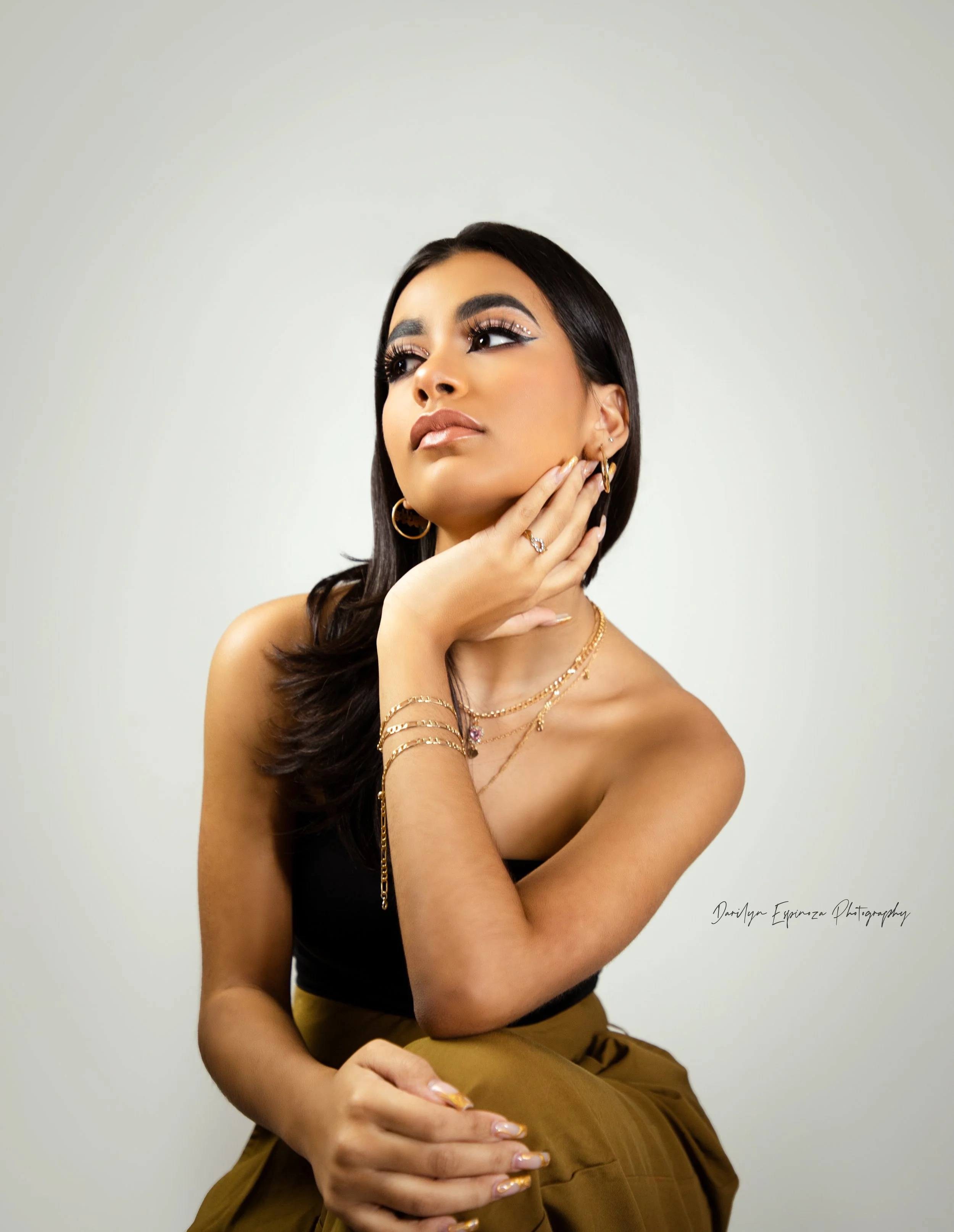 A woman with long dark hair and gold jewelry posing against a plain background.