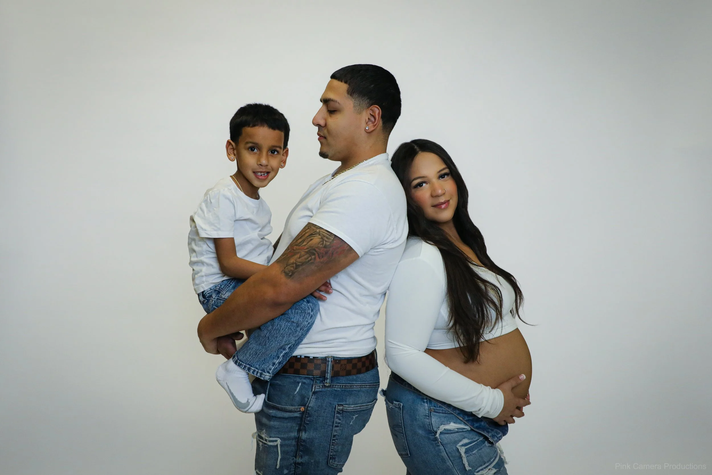 A family of three posing against a plain white background. The father is holding a young boy, and the pregnant mother is standing next to them, all dressed casually in white tops and jeans.