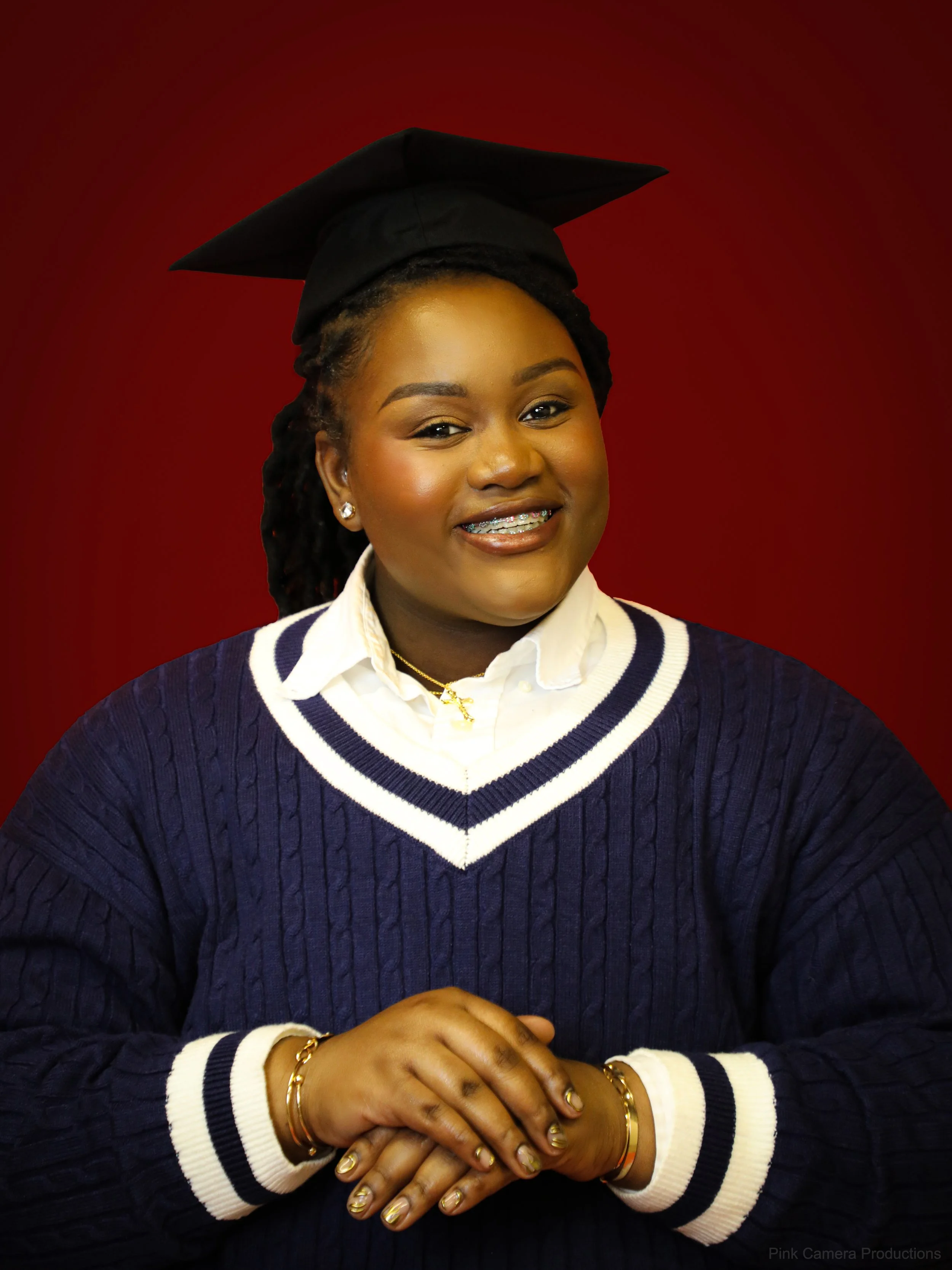 Young woman wearing a black graduation cap and gown, smiling, seated against a dark red background.