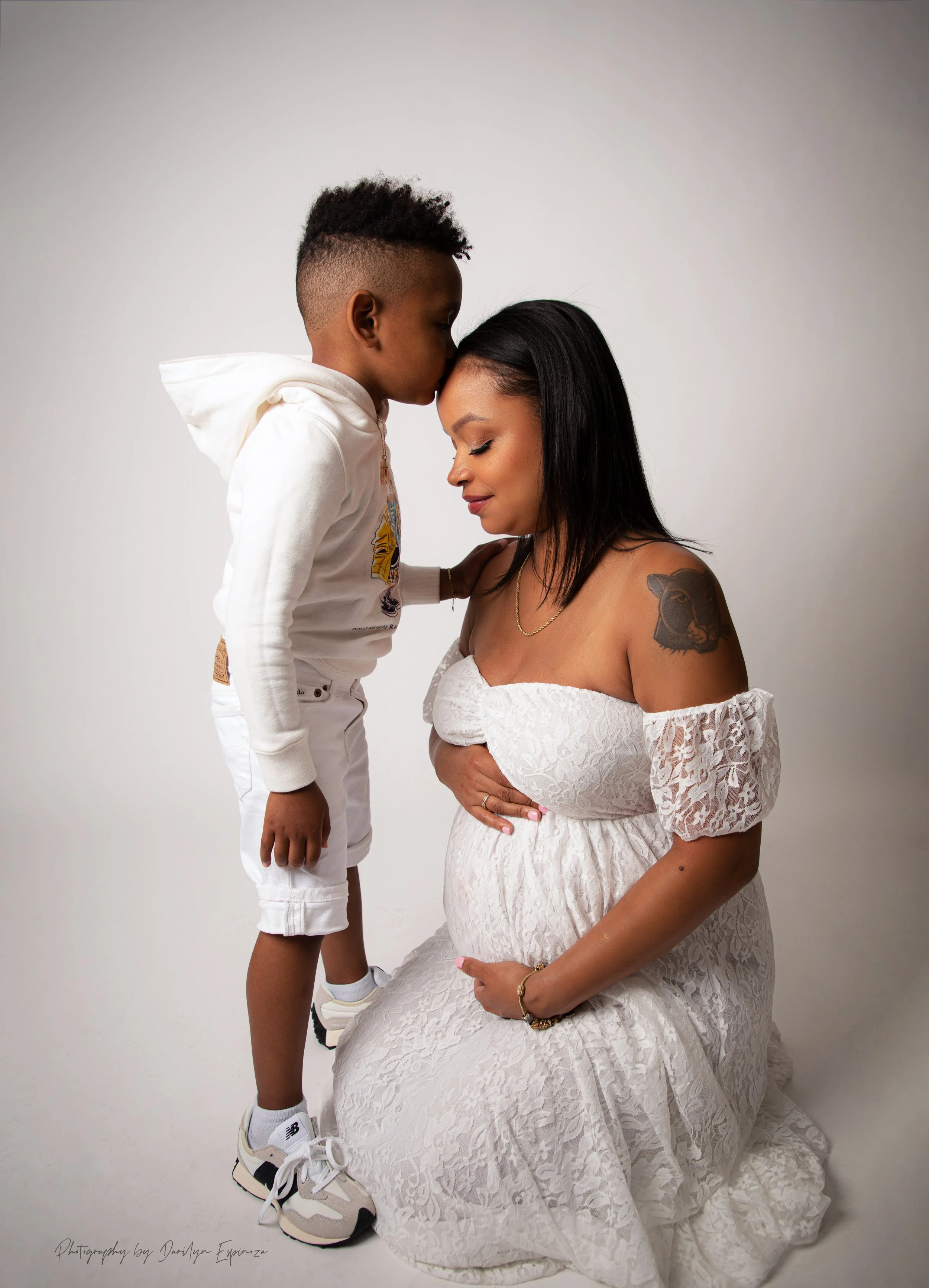 A woman in a white lace off-shoulder dress with a lion tattoo on her arm is kneeling and holding her pregnant belly, while a young boy in a white hoodie and shorts leans in to kiss her on the forehead in a studio with a plain white background.