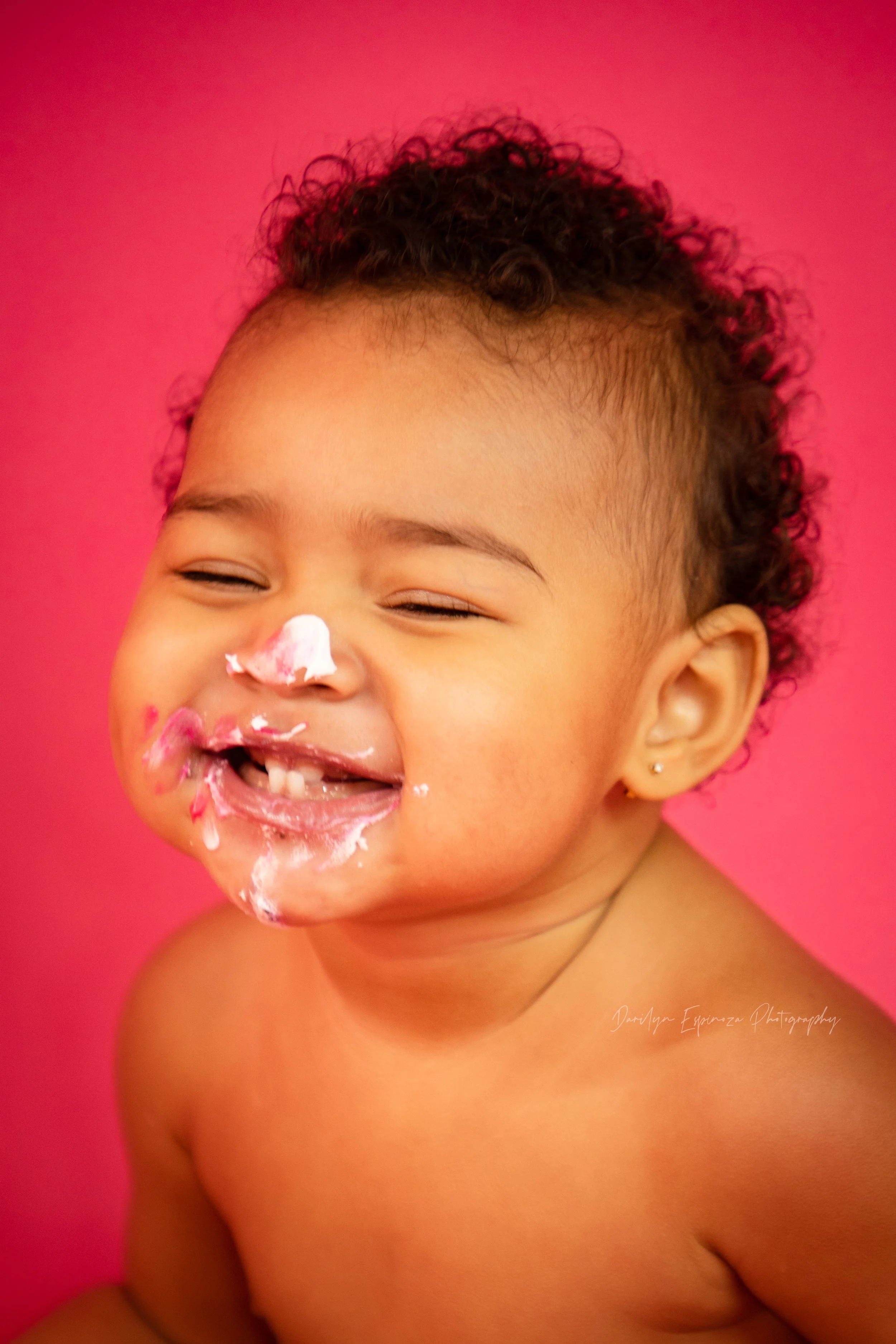 A young child with curly hair, smiling with eyes closed, covered in pink and white cake or frosting on face, against a pink background.