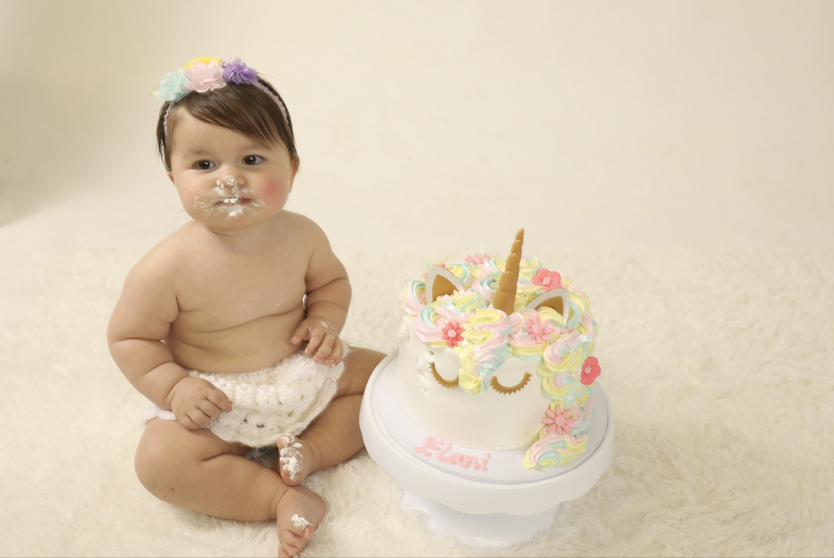 Baby girl with a flower headband sitting next to a unicorn-themed birthday cake.