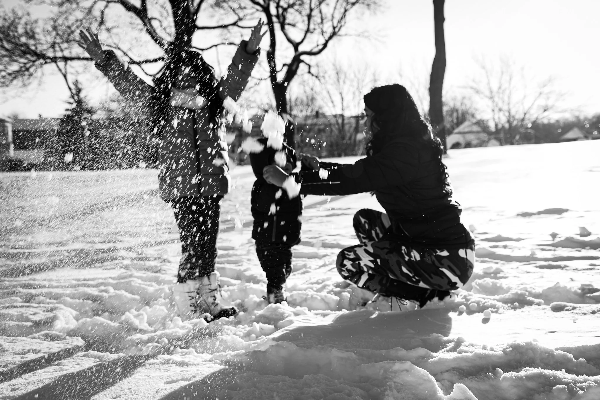 A woman and two children playing in the snow, with the woman kneeling and the children standing, throwing snow at each other in a winter outdoor setting with bare trees.