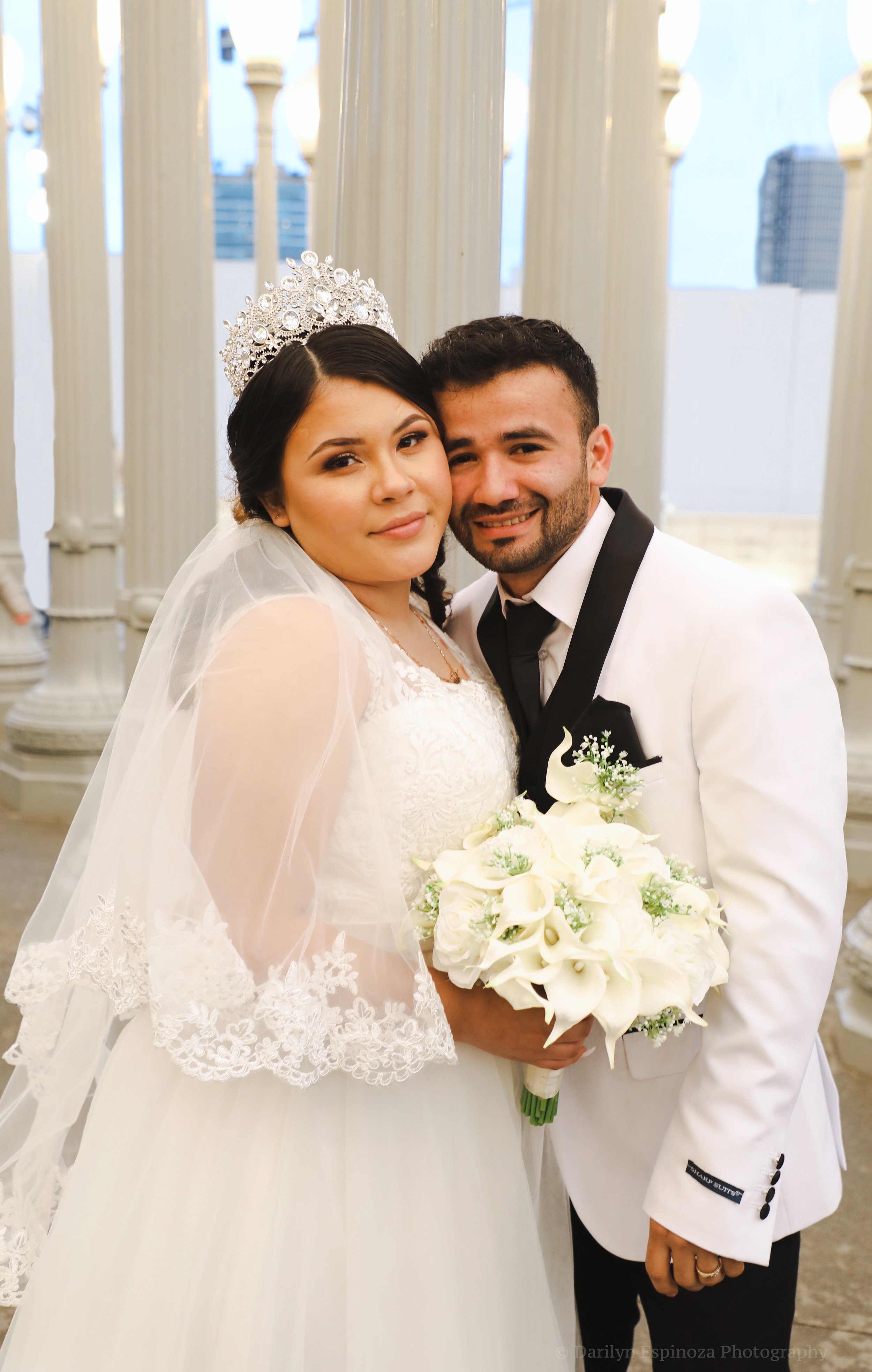 A bride in a white wedding dress and veil, wearing a tiara, holding a bouquet of white flowers, standing close to a groom in a white tuxedo with a black bow tie and black lapels, posing in front of white decorative columns outdoors.