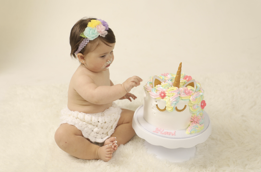 Baby girl celebrating her first birthday with a colorful unicorn-themed cake.