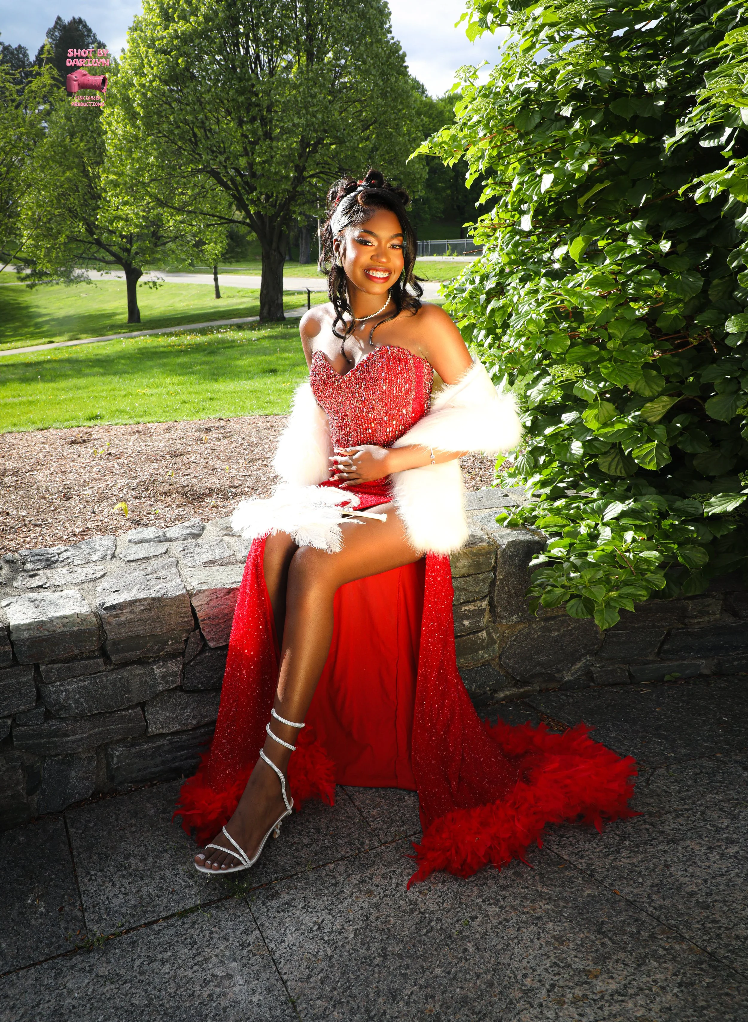 A woman in a red evening gown with a high slit, white fur shawl, and white high heels sitting on a stone wall in a park with green trees and grass, smiling at the camera.