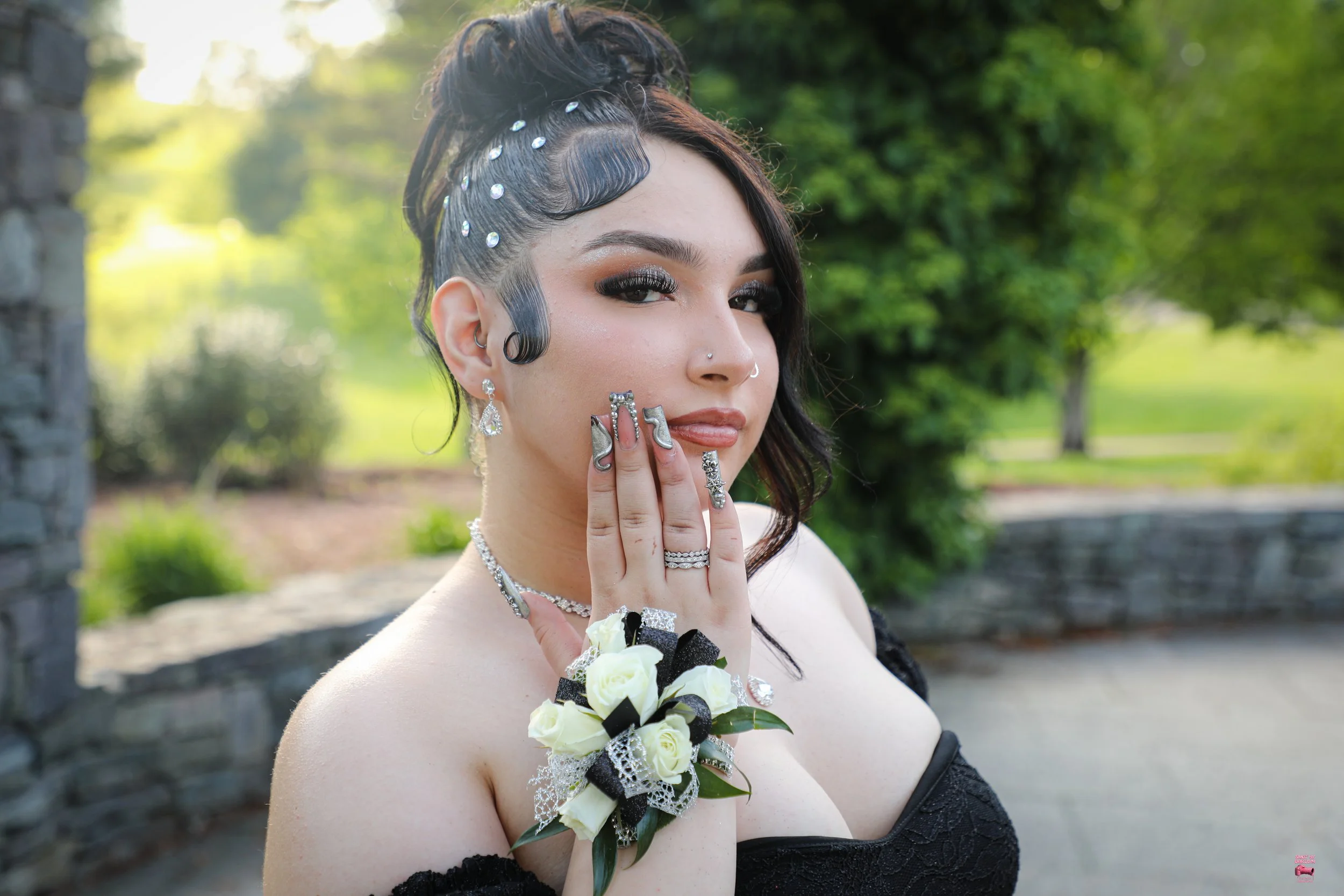 A woman dressed in black with jewelry and a corsage, standing outdoors with trees and stone structures in the background.