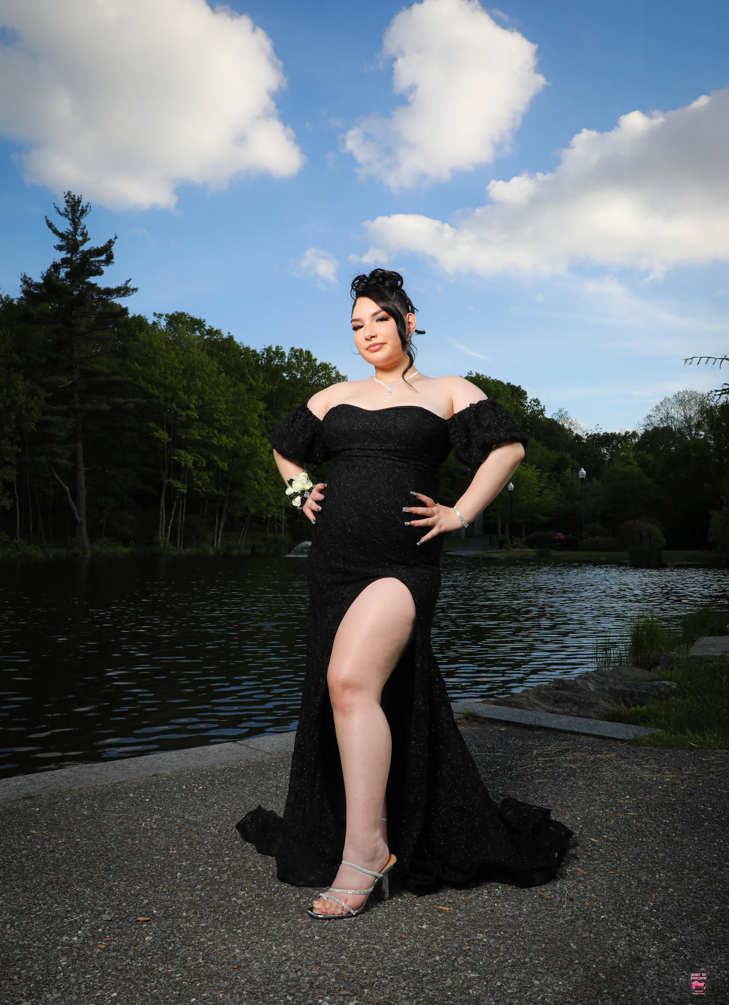 A woman in a black evening gown with a high slit, standing by a lake during the day, with trees and a blue sky with clouds in the background.