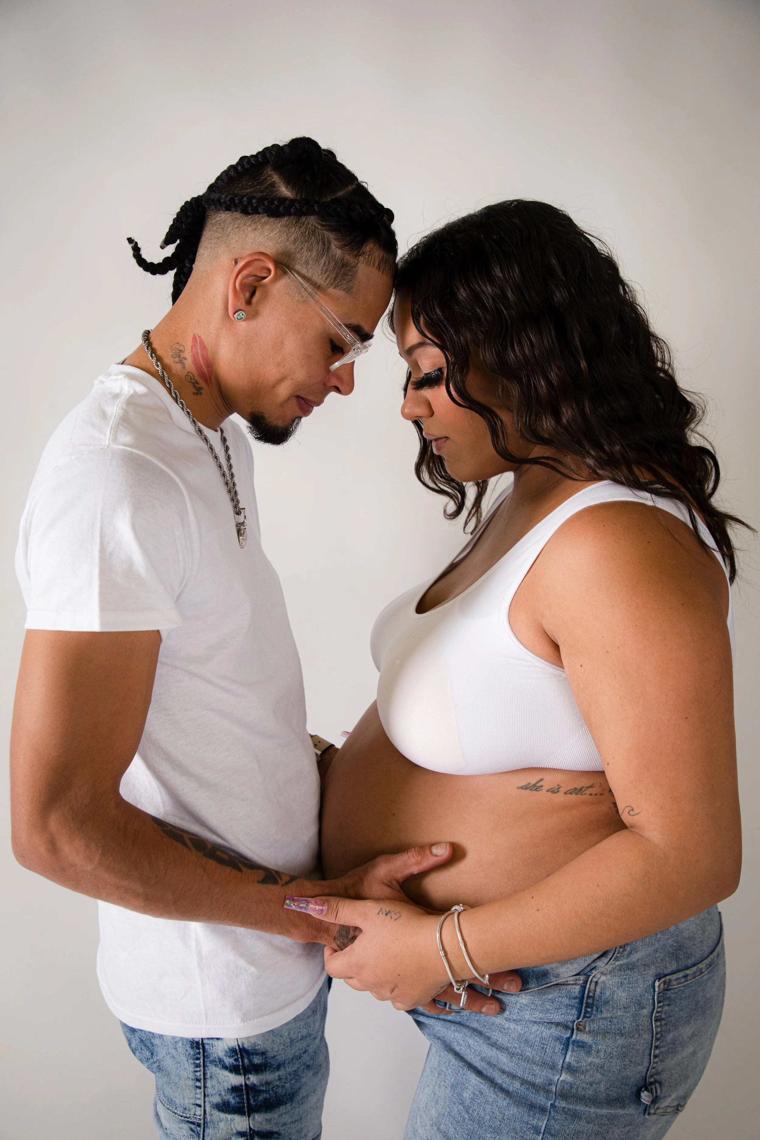 A couple showing their pregnant bellies, with foreheads touching and hands on the belly, against a plain background.