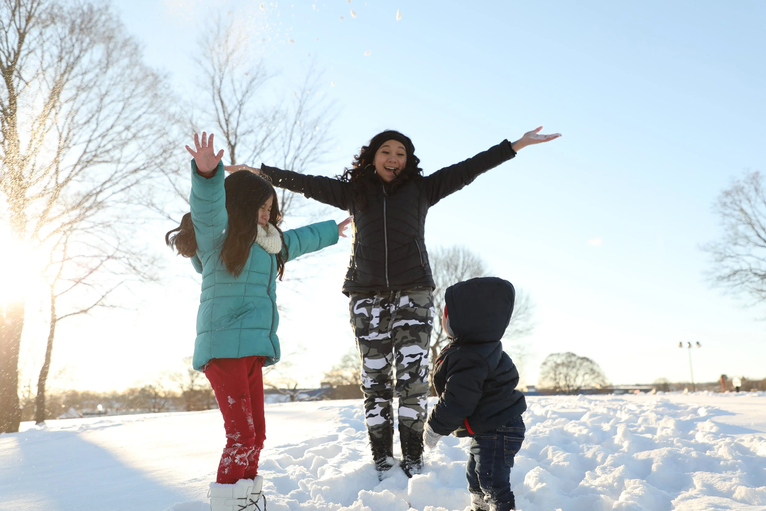 A woman and two children playing in the snow in a park during winter, with leafless trees and a clear sky in the background.