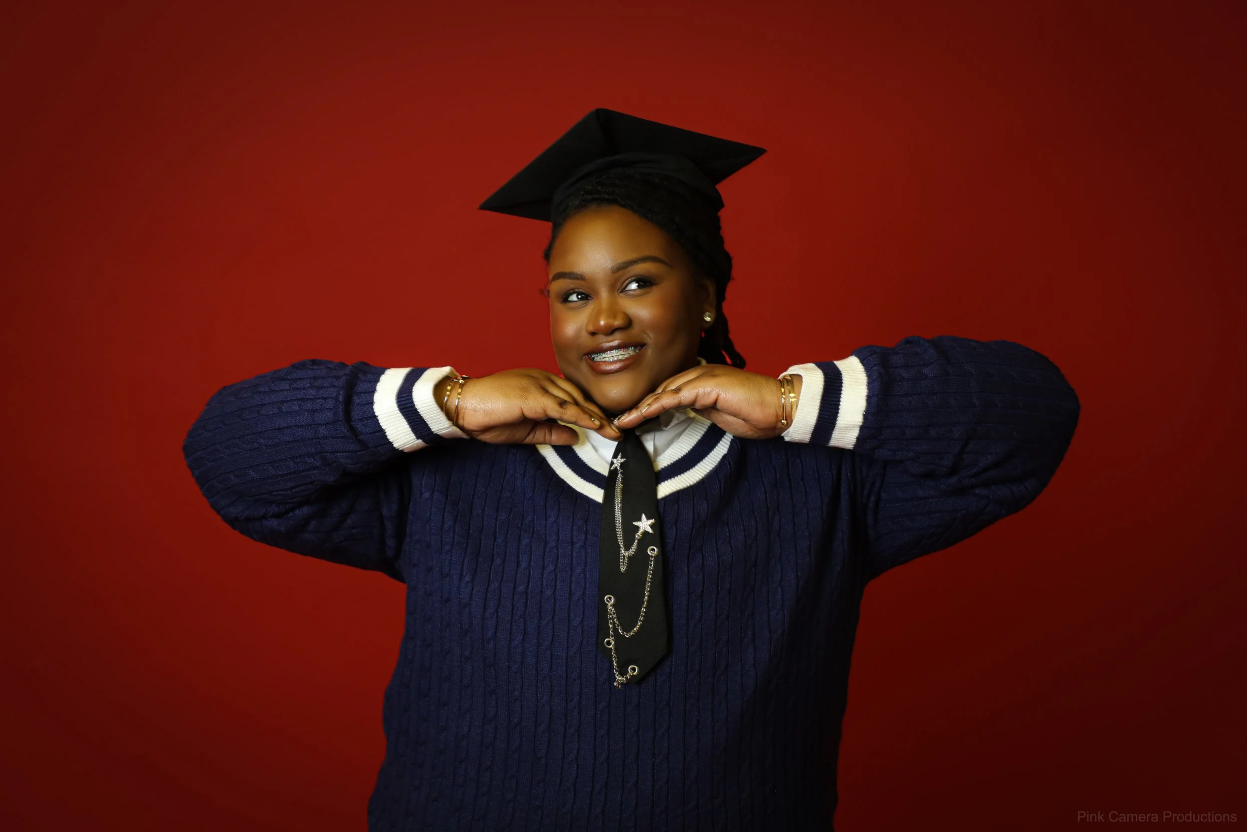 Young woman in a graduation cap and navy sweater posing with hands under chin against a red background.