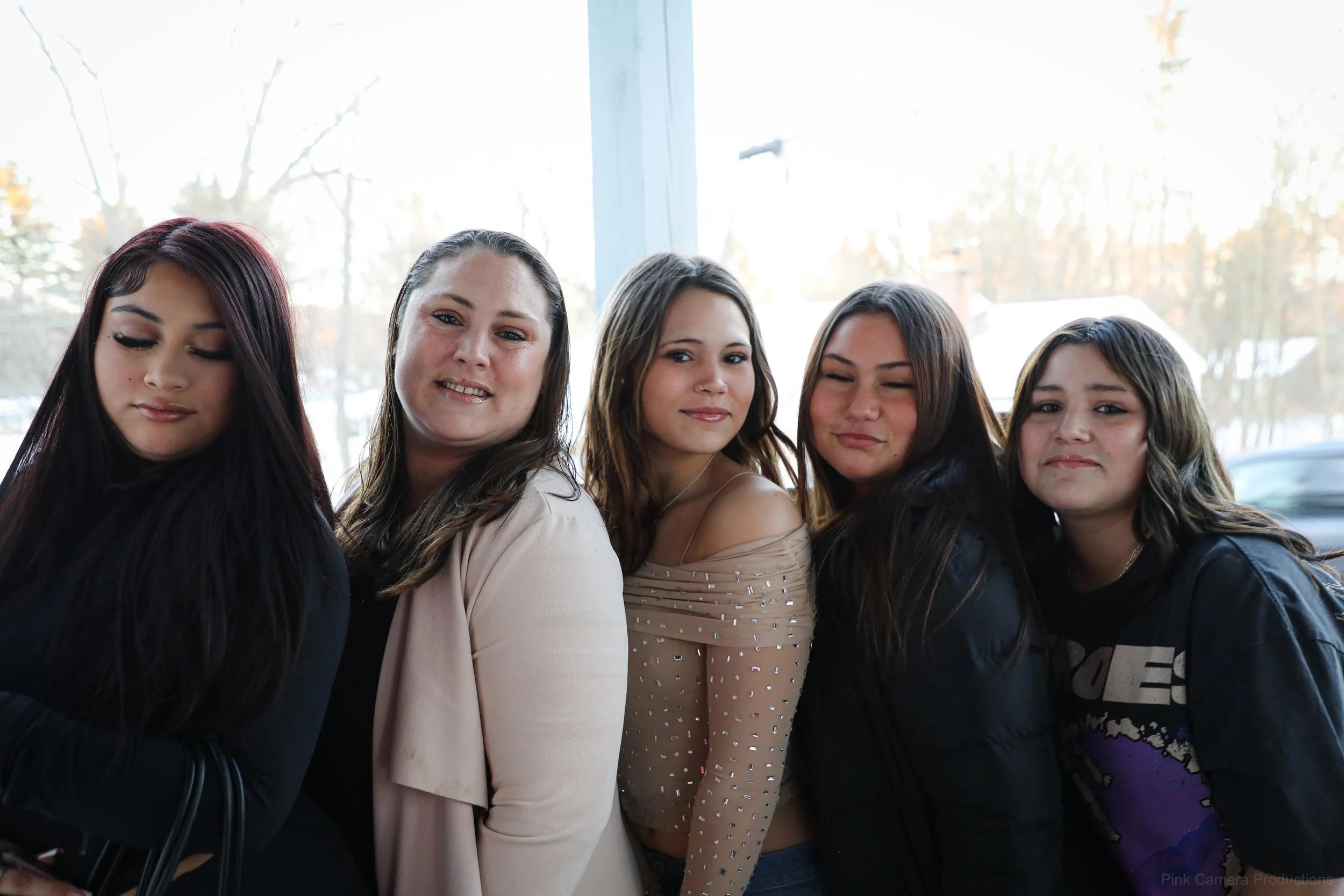 Five young women standing close together inside, with a large window behind them showing a snowy outdoor scene.