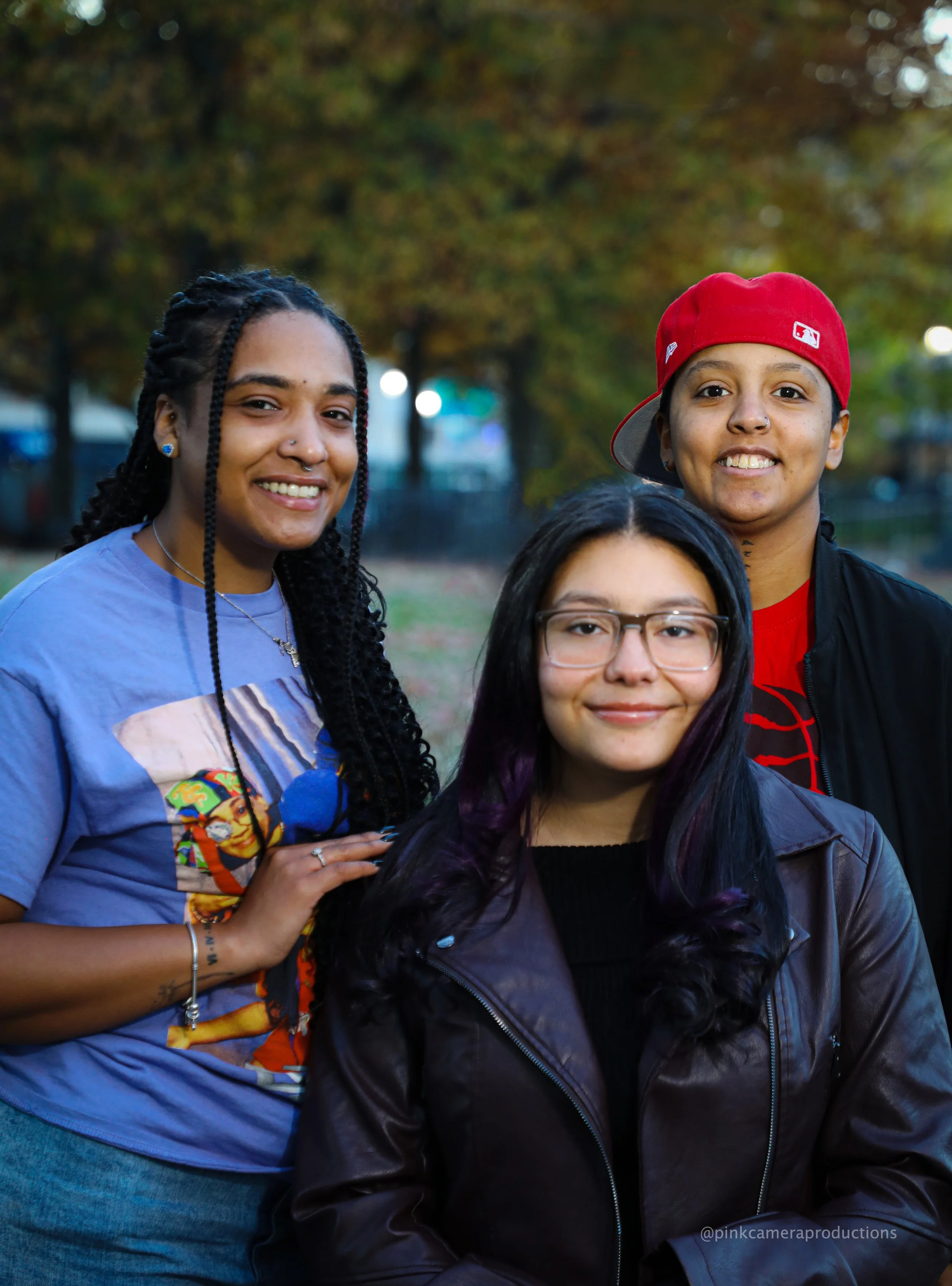 Three young women standing together outdoors in a park with autumn foliage in the background, smiling at the camera.
