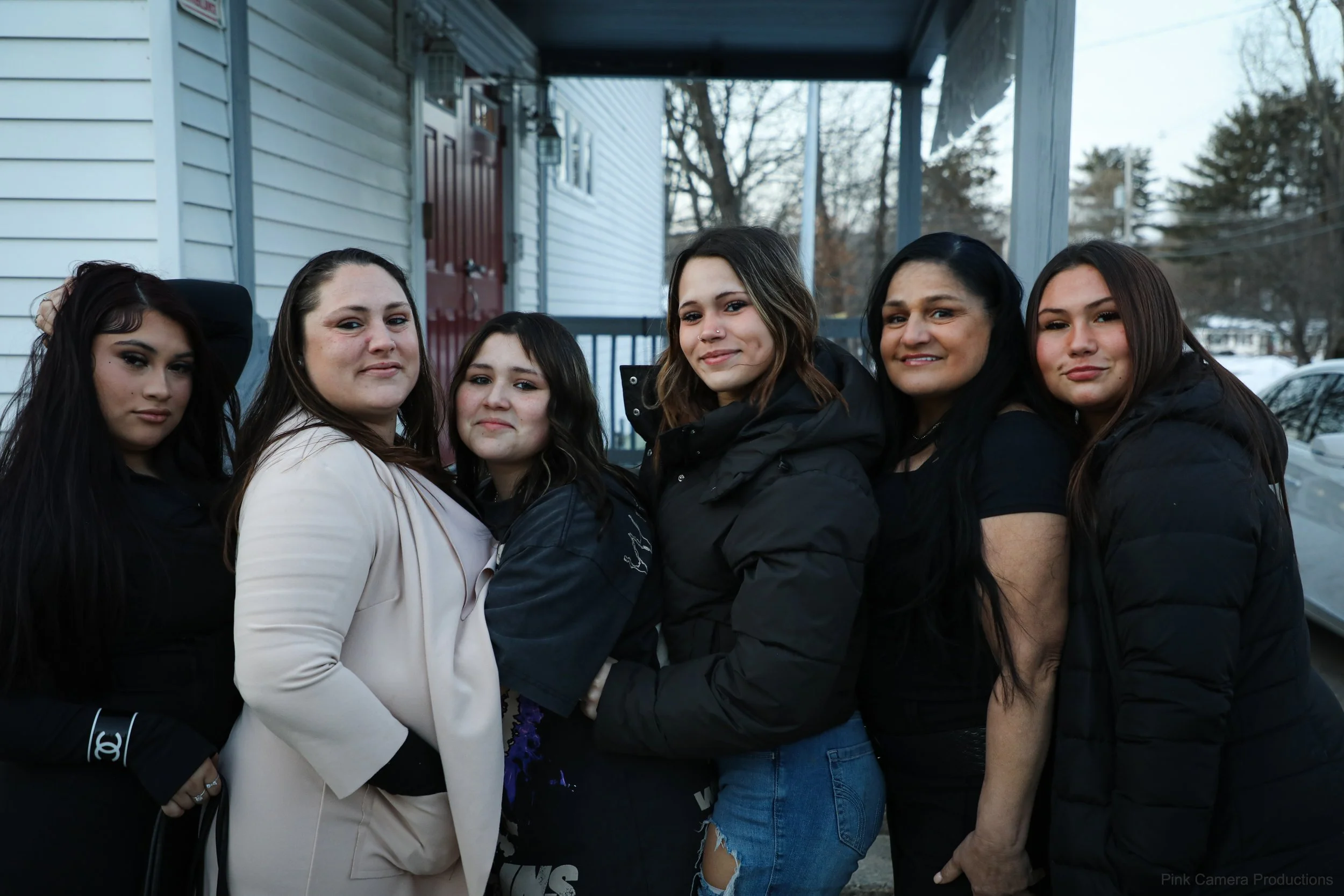 A group of six women standing closely together outside on a porch, with a house and trees in the background. They are dressed in casual winter clothing and appear to be posing for the photo.