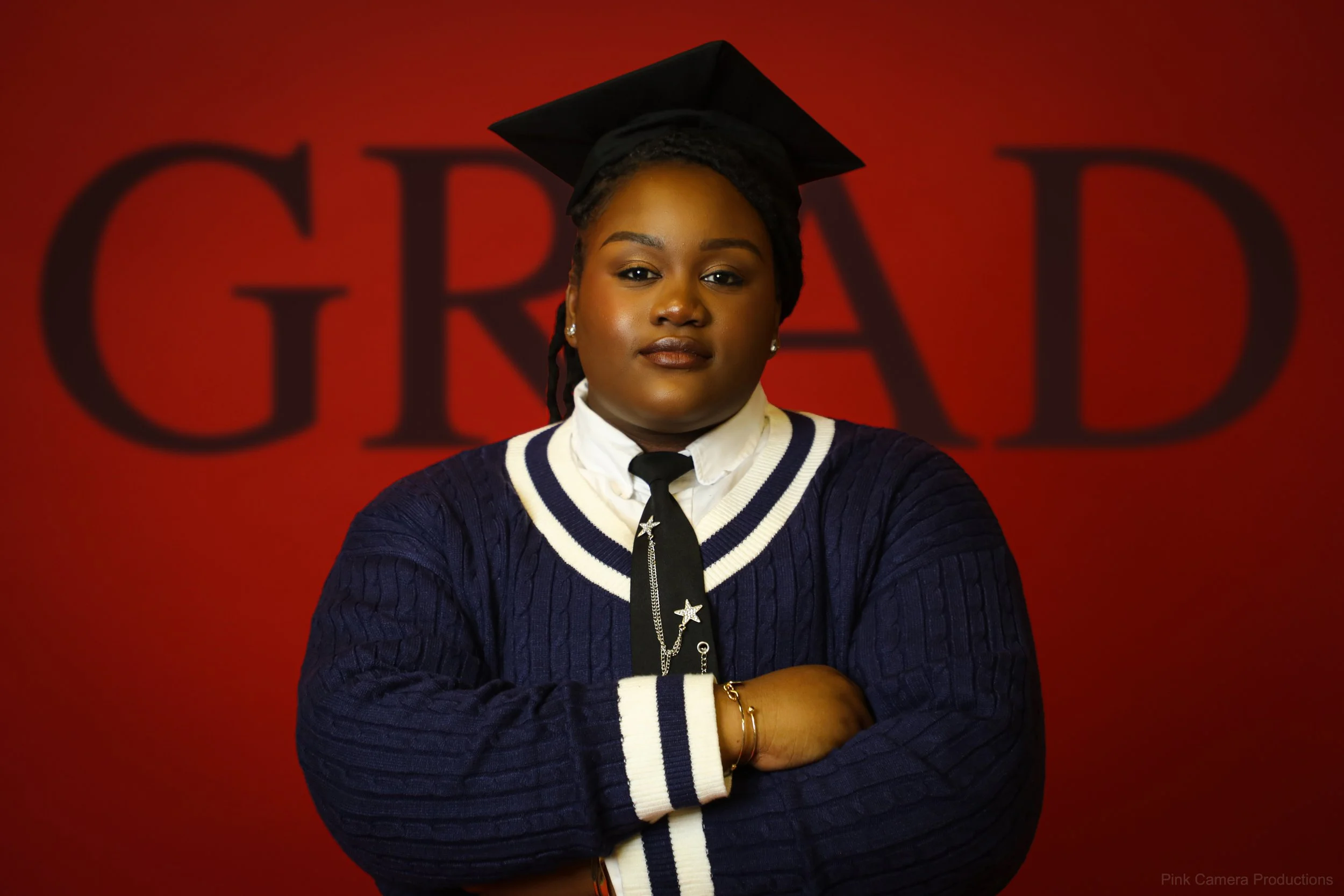 A young woman in a graduation cap and gown with crossed arms, standing in front of a red background with the word 'GRAD' partially visible.