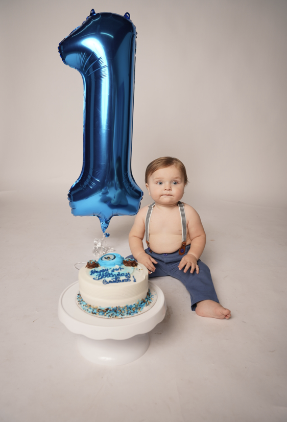 A baby sitting next to a birthday cake with a blue number one balloon behind them.