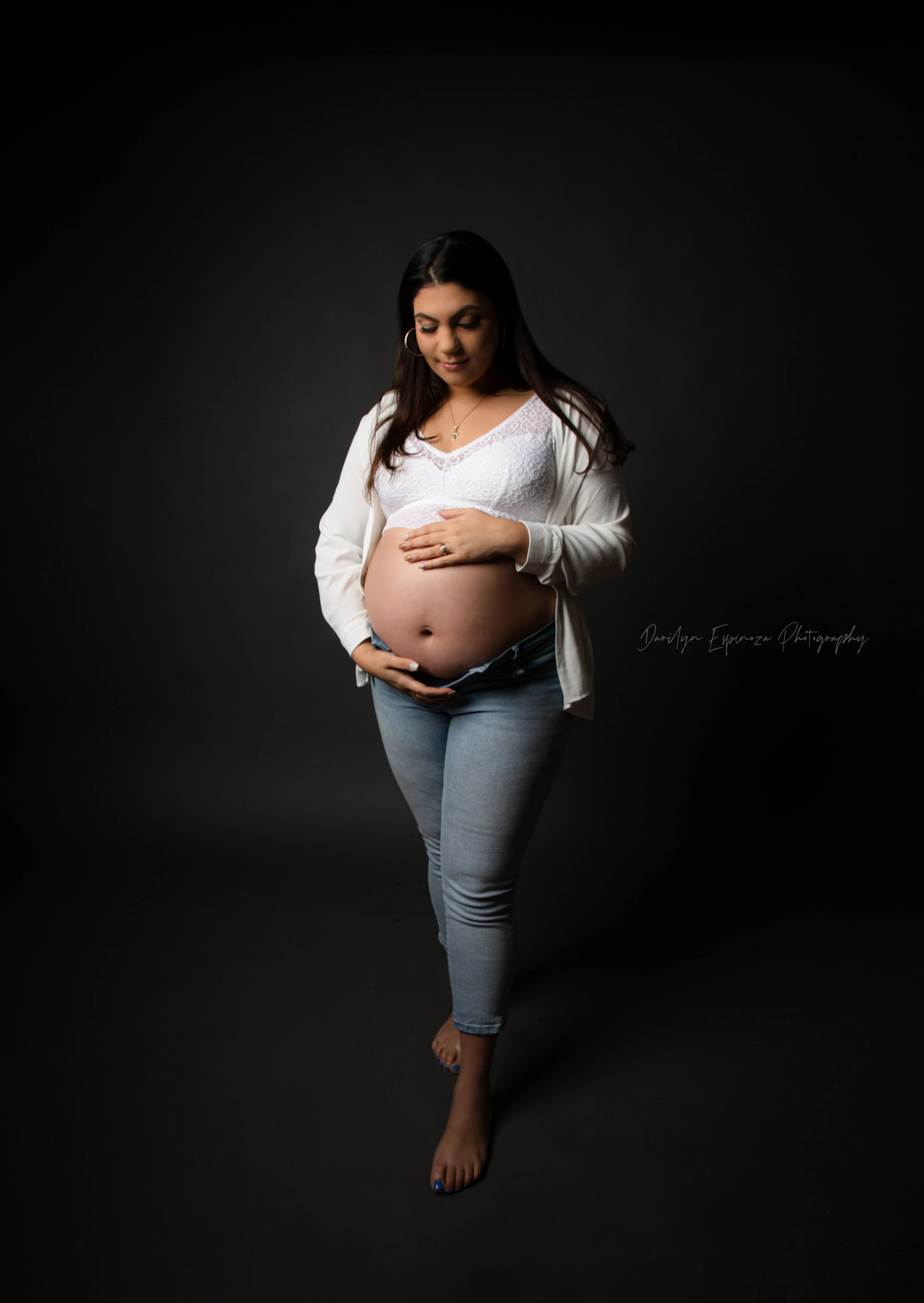Pregnant woman with long dark hair, holding her belly, wearing a white top, light blue jeans, and a white jacket, standing barefoot against a dark background.