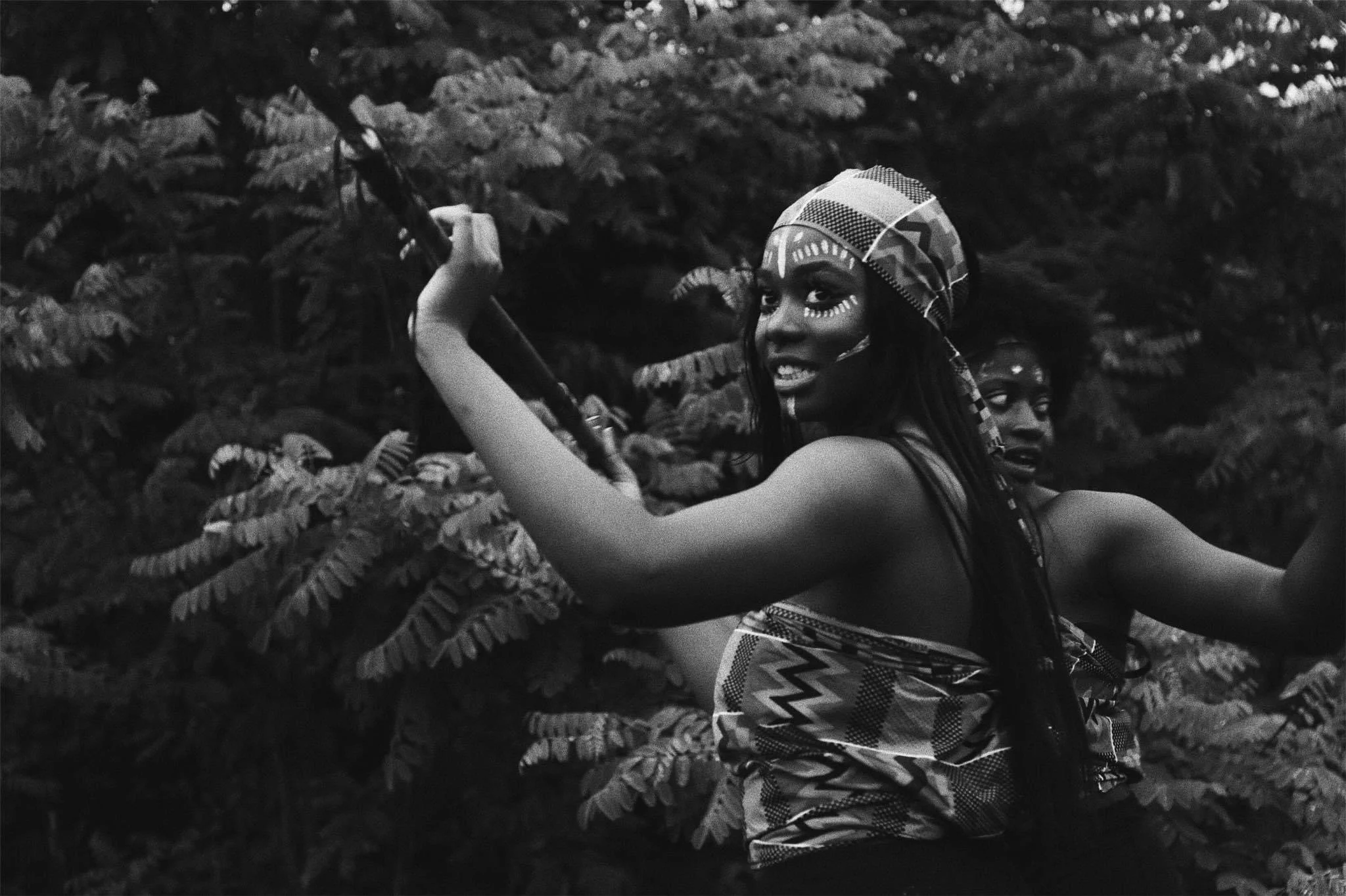 Two women in traditional Maasai attire dancing among lush foliage, one smiling and wearing a headscarf, in a black and white photograph.
