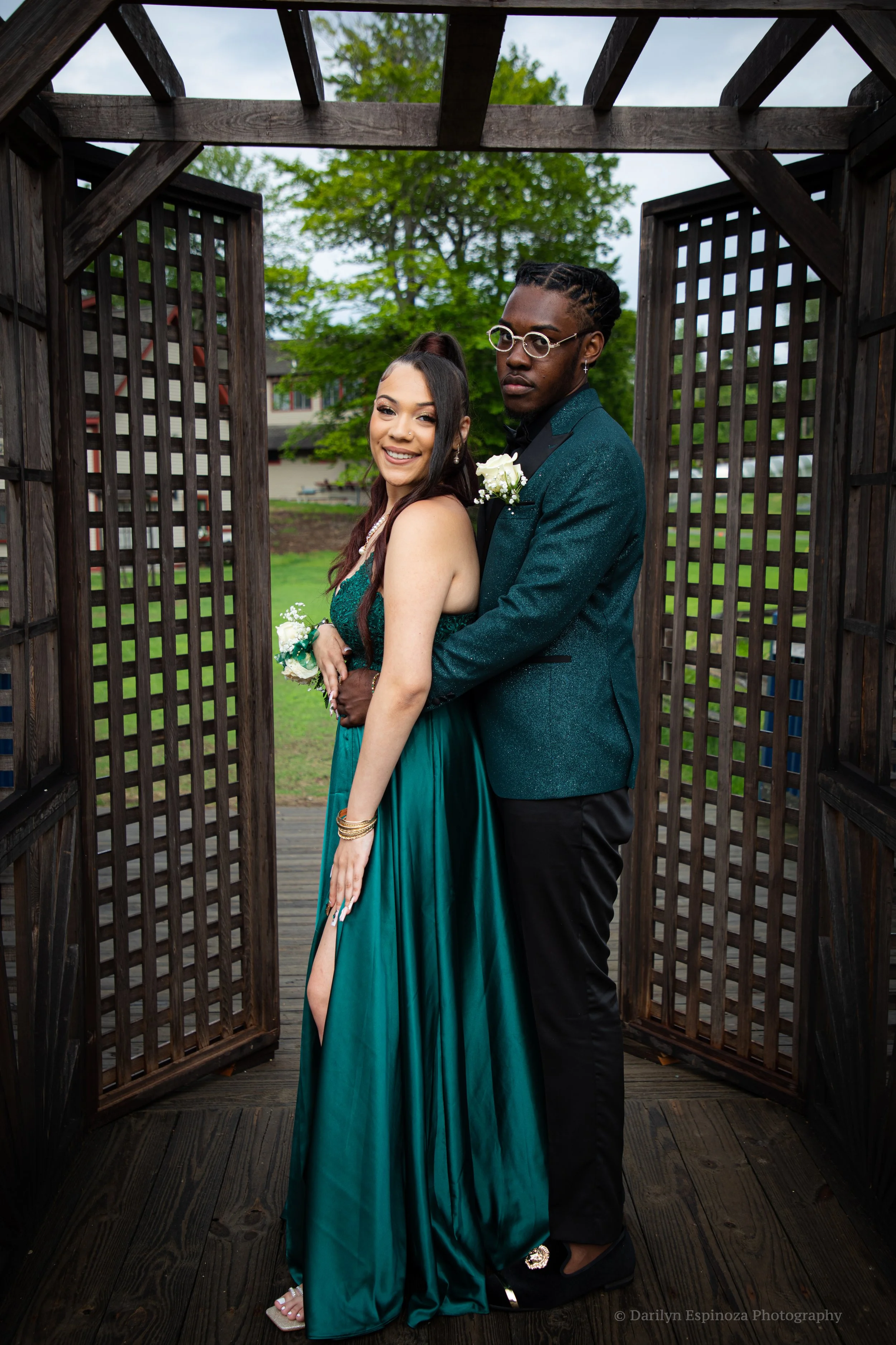 A young couple dressed in formal attire, standing close together in a wooden gazebo with a lush green lawn and trees in the background. The woman is smiling, wearing a teal dress with a high slit, and the man is wearing a dark teal blazer and black t