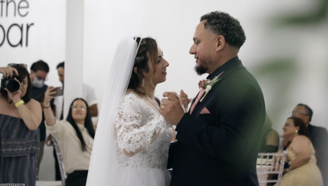 A bride and groom are dancing during their wedding ceremony, surrounded by seated guests and photographers.