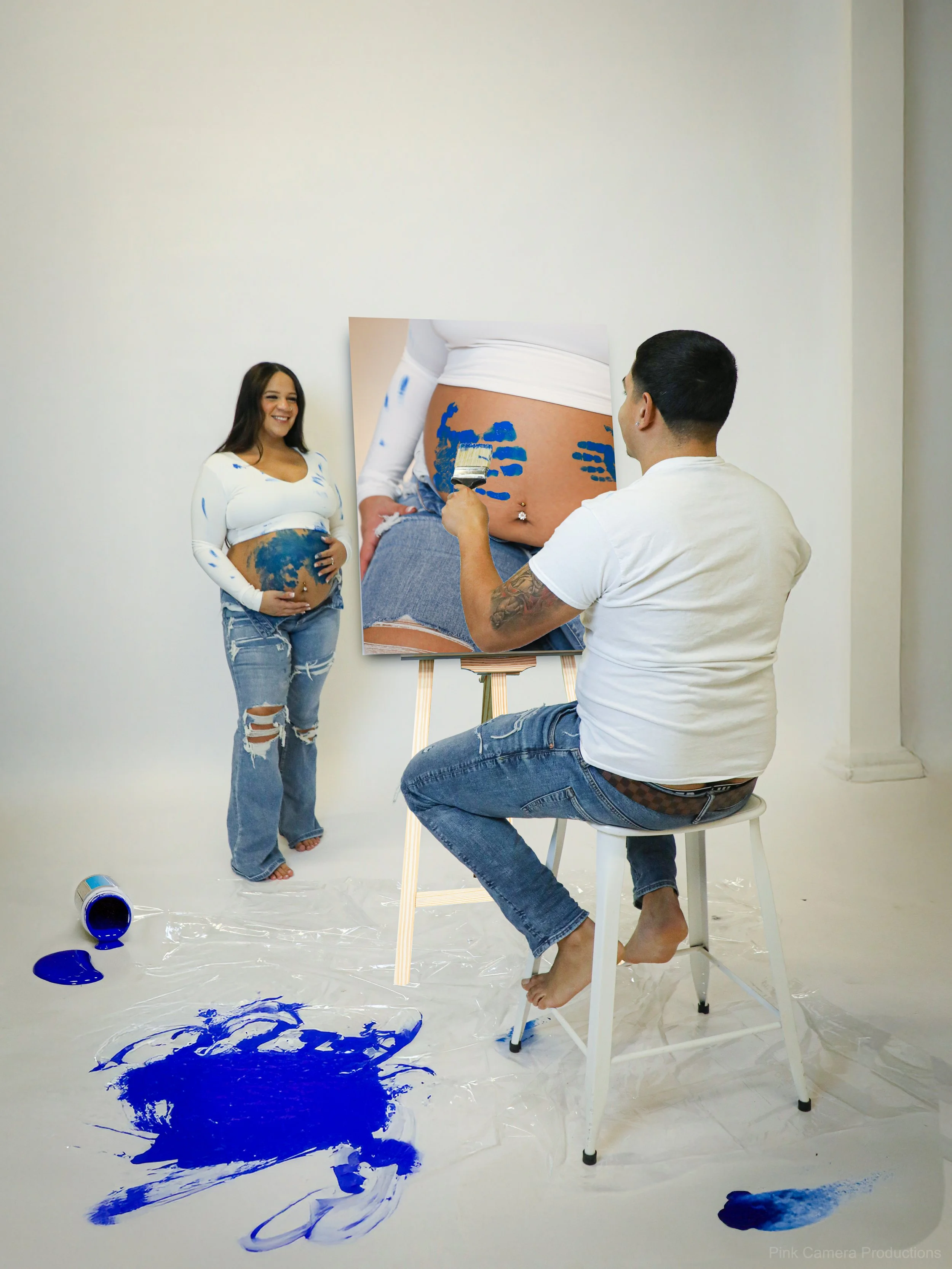 A woman with a pregnancy belly painted with blue paint, standing smiling in a white long sleeve shirt and ripped jeans. A man is seated on a white stool, painting her belly on a canvas that features a close-up scene of her belly with blue paint and a