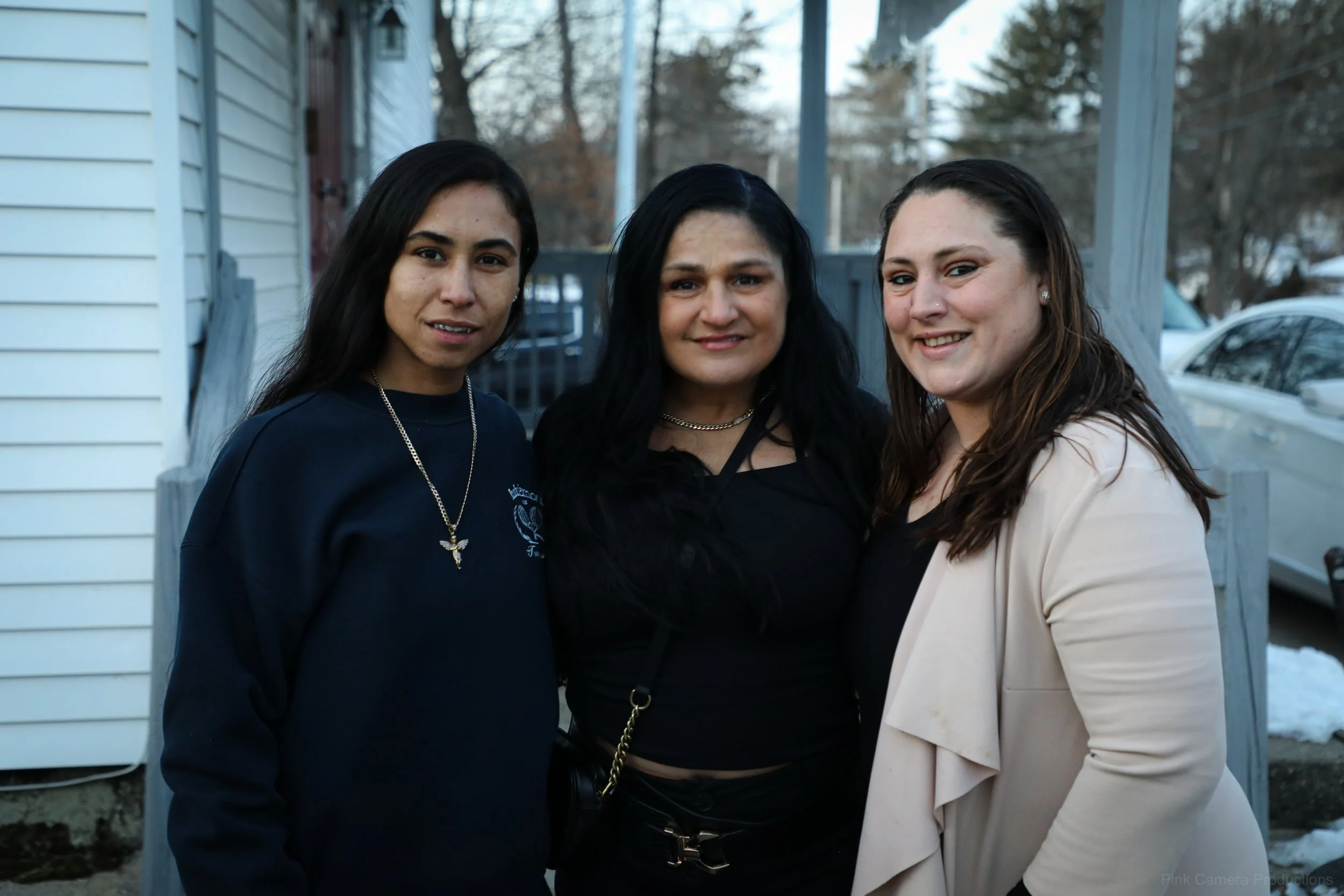 Three women standing outdoors in front of a house, smiling at the camera, with trees and cars in the background.