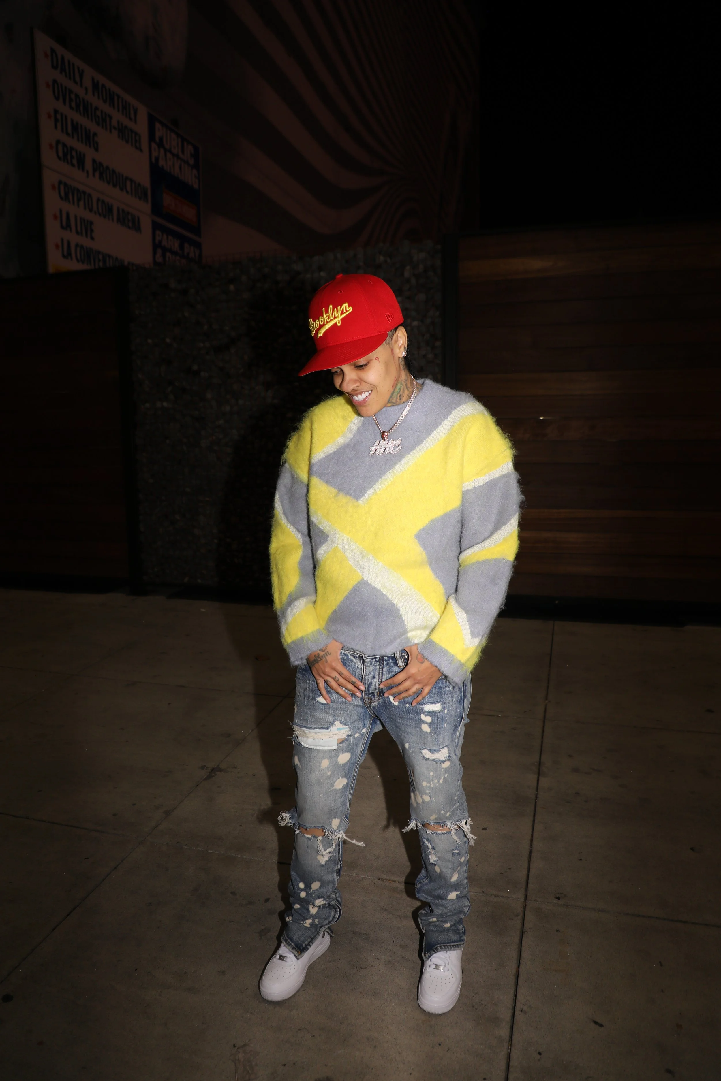 A woman standing outdoors at night, wearing a yellow and gray sweater, ripped jeans, white sneakers, and a red cap with 'Roselwyn' embroidered on it, smiling and looking downward.