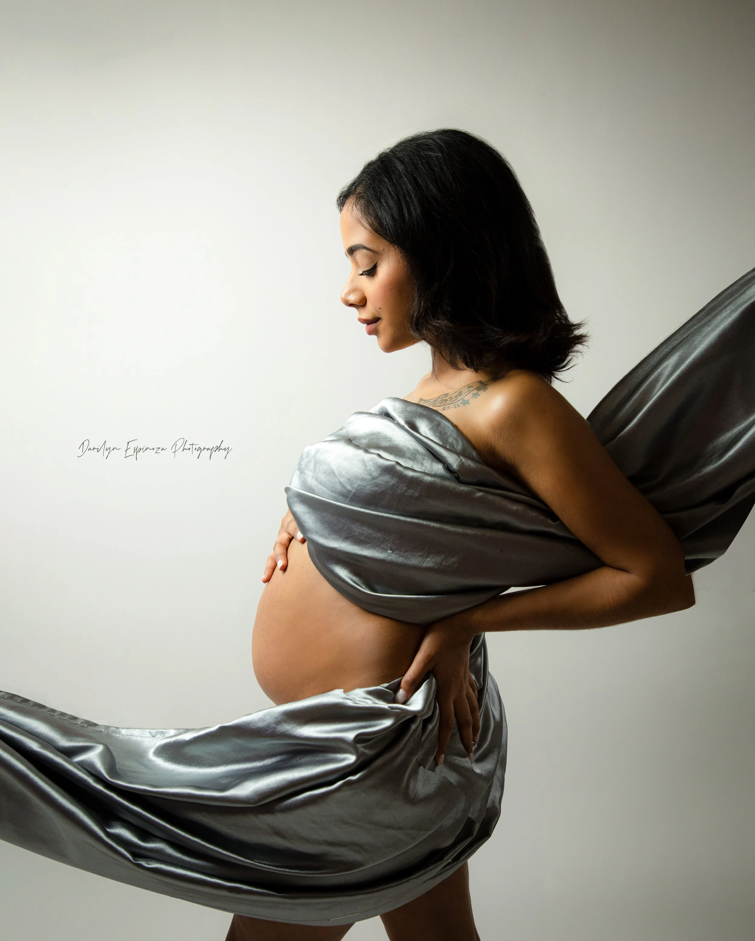 Pregnant woman with dark hair posing with silver satin fabric draped around her, standing in profile against a plain light background.