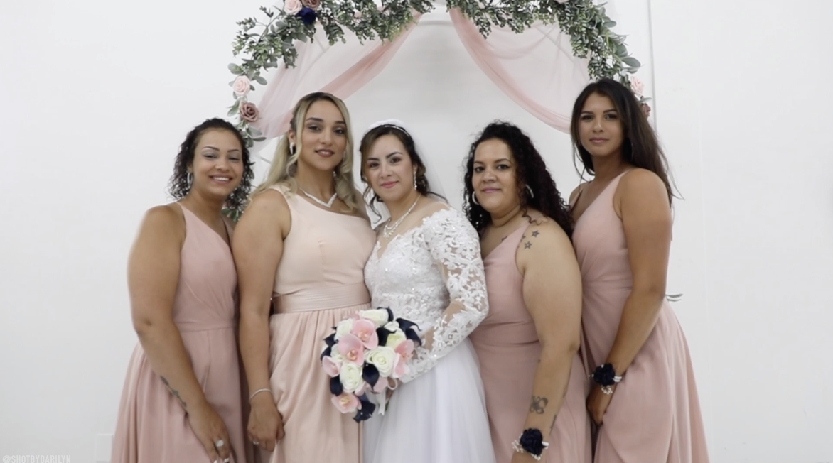 Bride in white wedding dress holding a bouquet, surrounded by five bridesmaids in pink dresses, in front of a floral arch with pink fabric