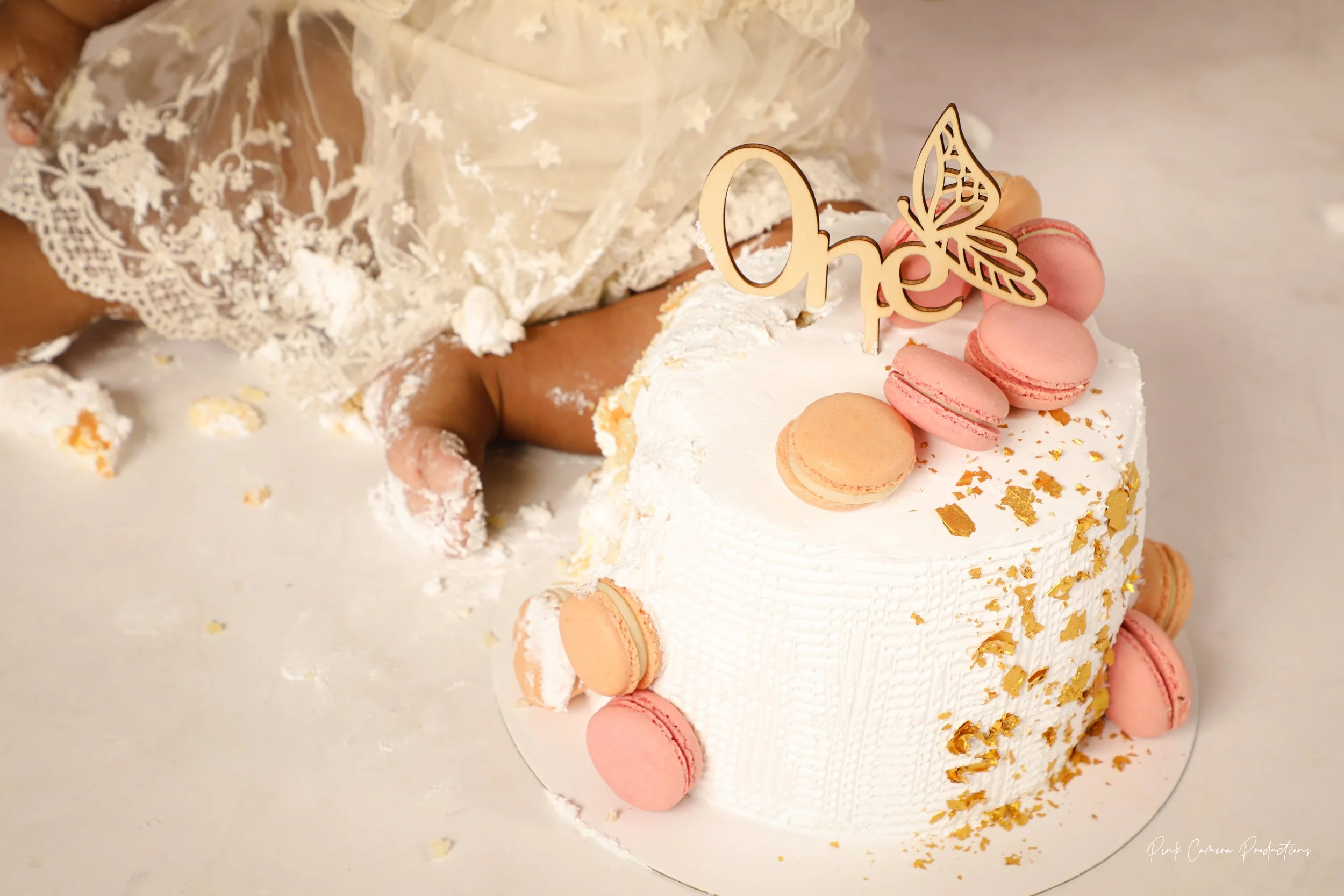 Close-up of a white birthday cake with pink macarons and gold leaf decorations, topped with a wooden "One" cake topper with a butterfly design, with a child's hand in a lace dress reaching for the cake.