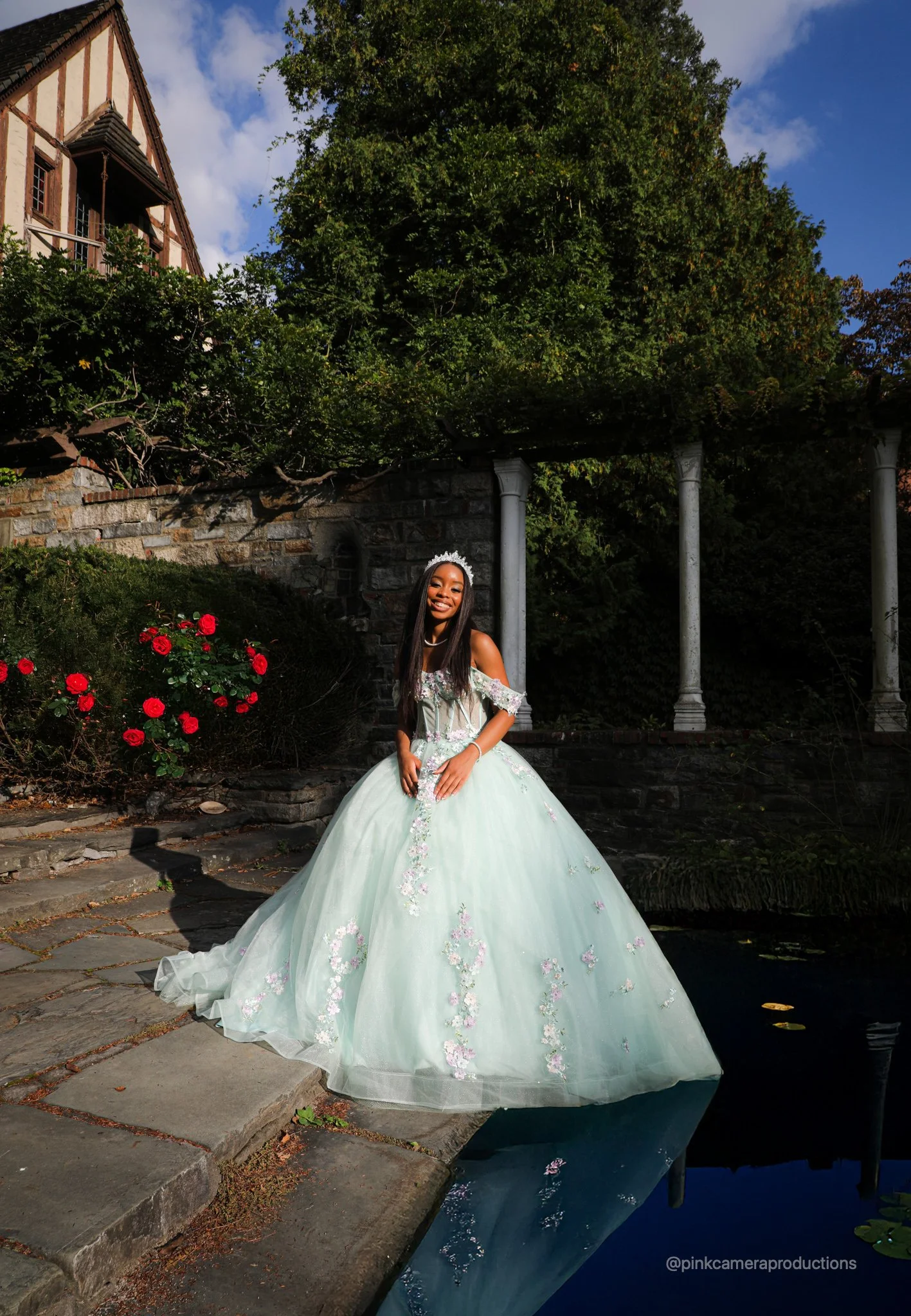 A young woman in a light green, floral quinceañera gown with off-shoulder sleeves and a tiara stands by a pond, smiling, outdoors with a garden, roses, stone pathway, and columns in the background.