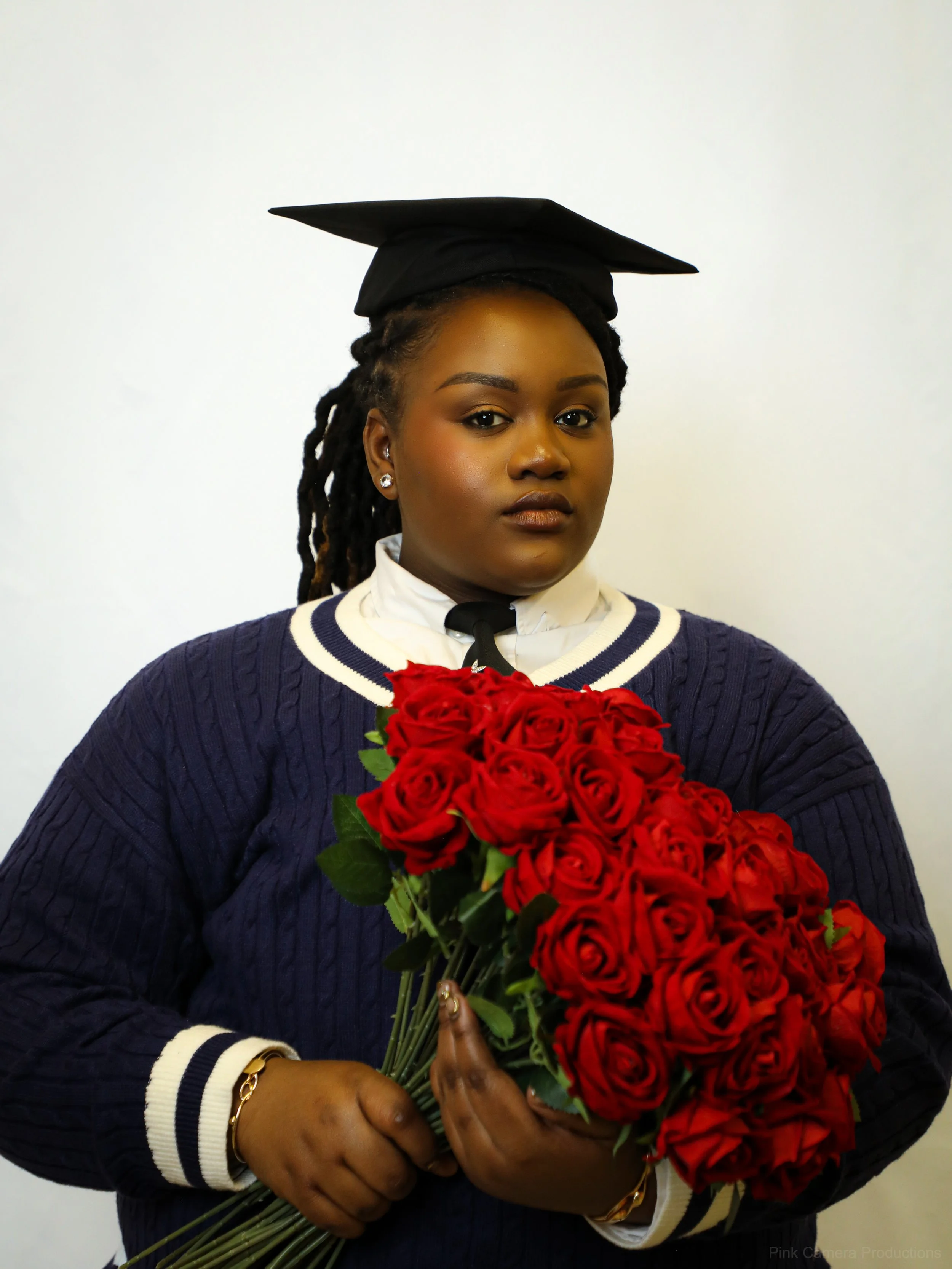 A woman with dark skin and braids wearing a graduation cap and gown holding a large bouquet of red roses.