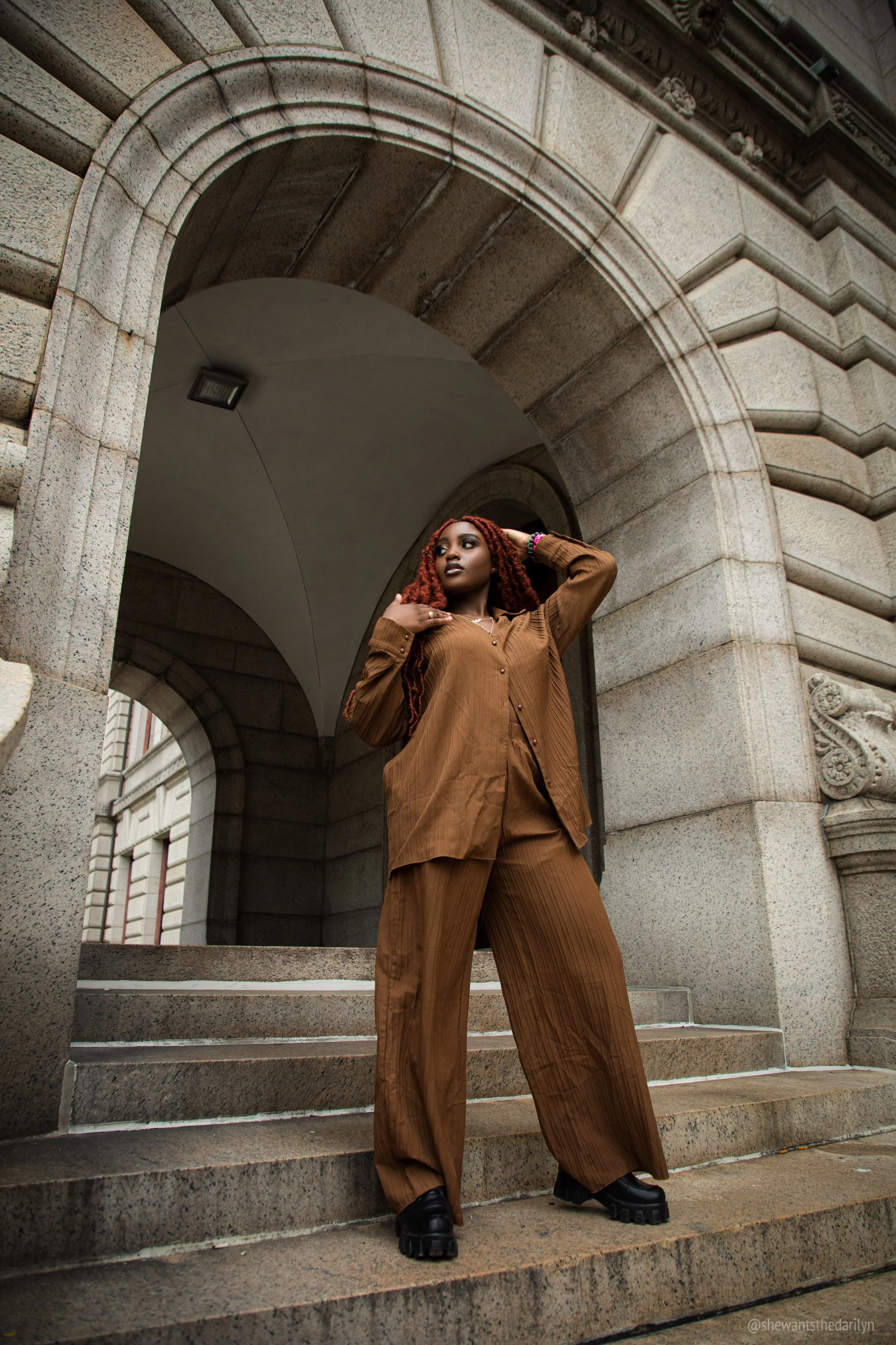 A woman with red dreadlocks wearing a brown suit with wide-leg pants and a jacket, standing on stone steps beneath an archway of a historic building.
