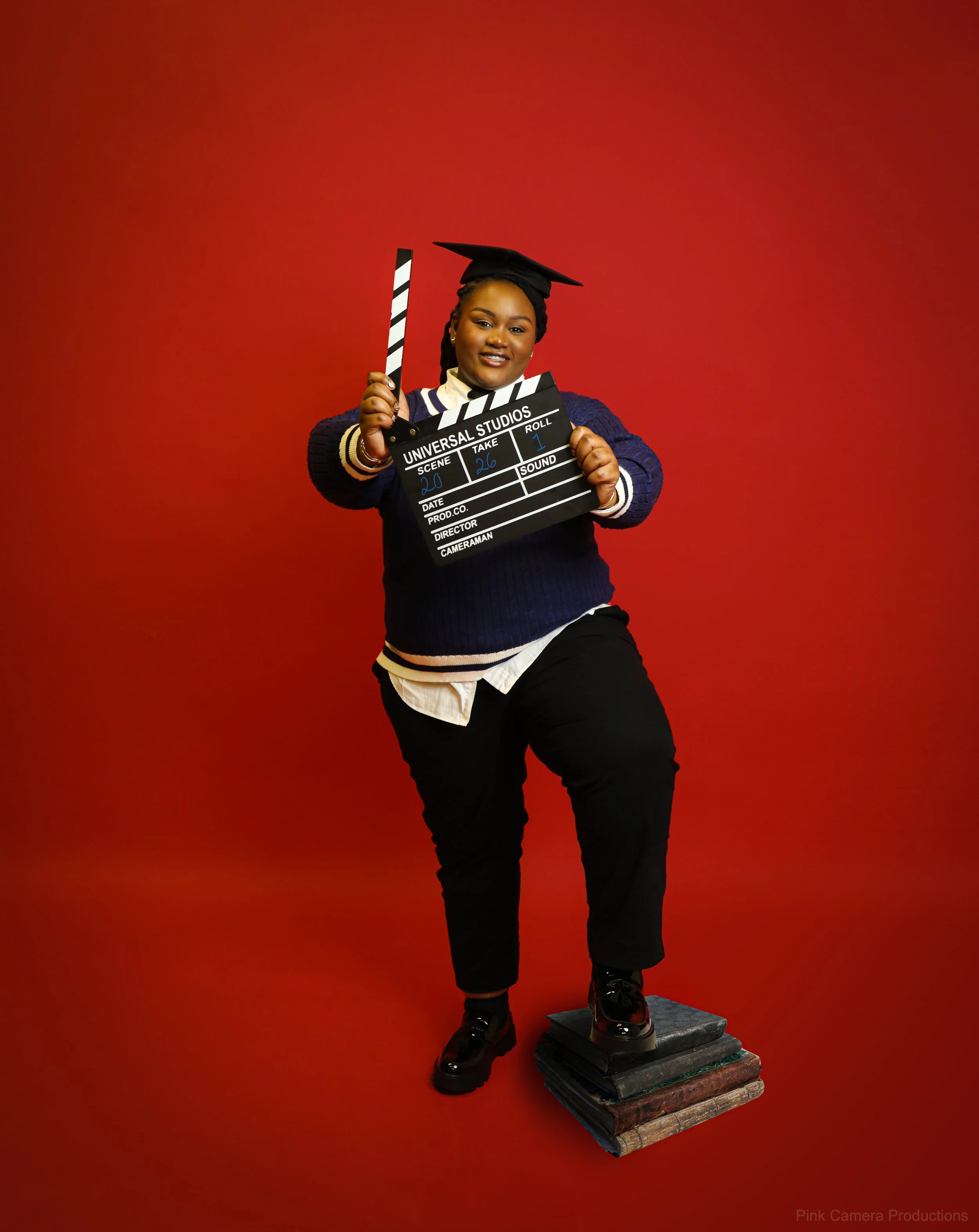 A woman in a graduation cap and casual attire holding a film clapperboard, standing on a stack of old books against a red background.