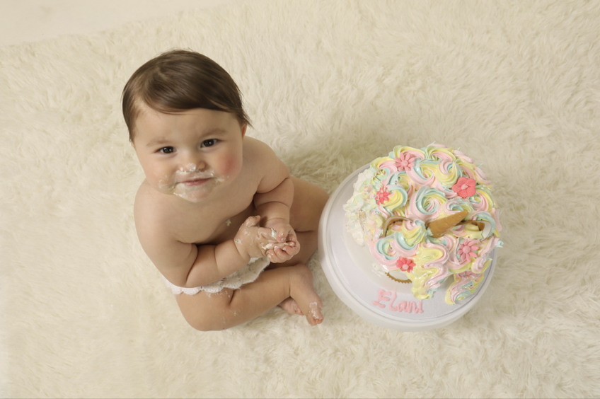 A baby with cake frosting on their face and hands, sitting on a plush light-colored carpet next to a colorful birthday cake with pastel swirls and a unicorn-shaped decoration.