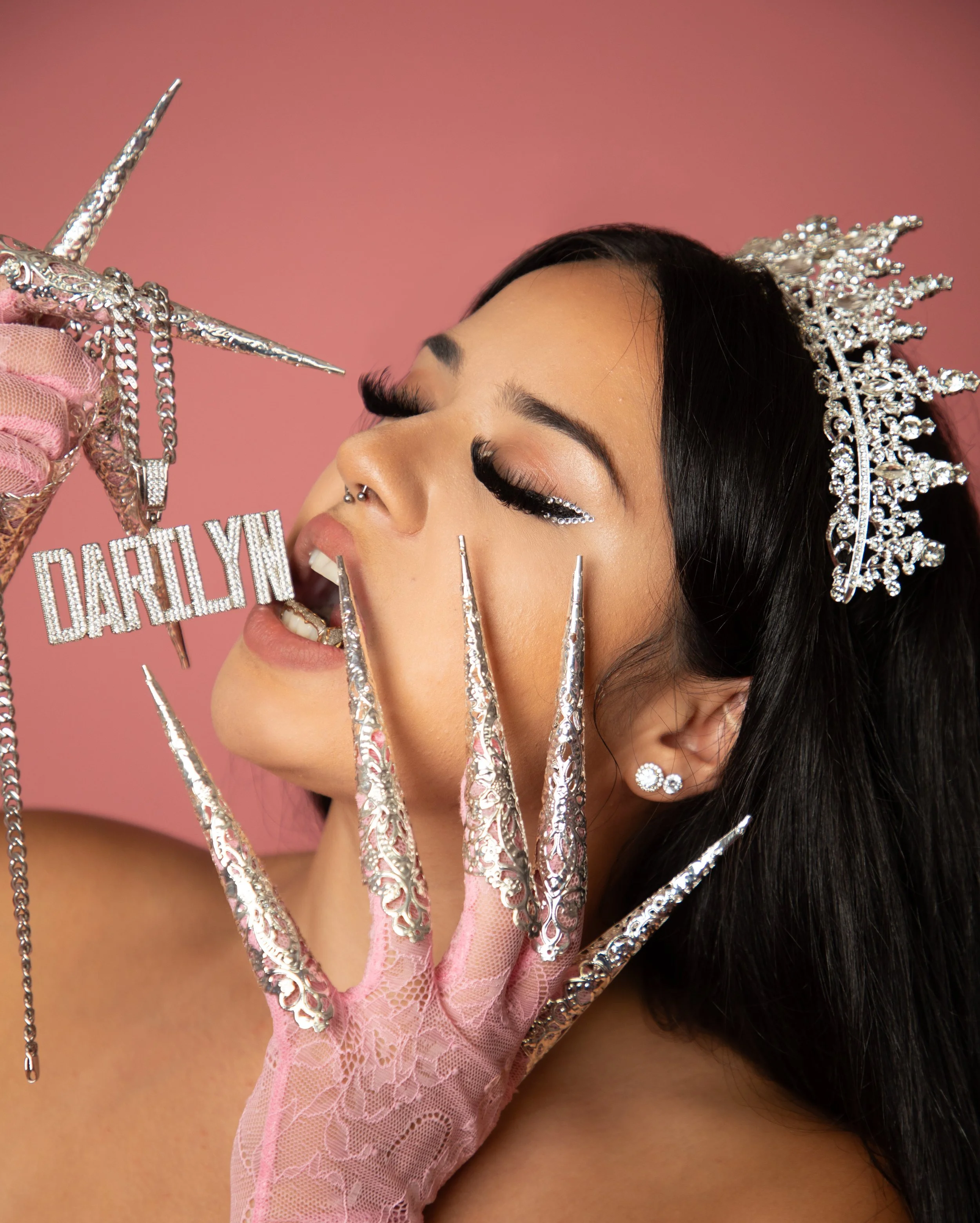A woman with long dark hair and dramatic makeup, wearing a sparkly tiara and earrings, holding a diamond necklace with the name "Darilyn" in rhinestones. She has long, ornate, silver nails and is dressed in pink lace gloves against a pink background.