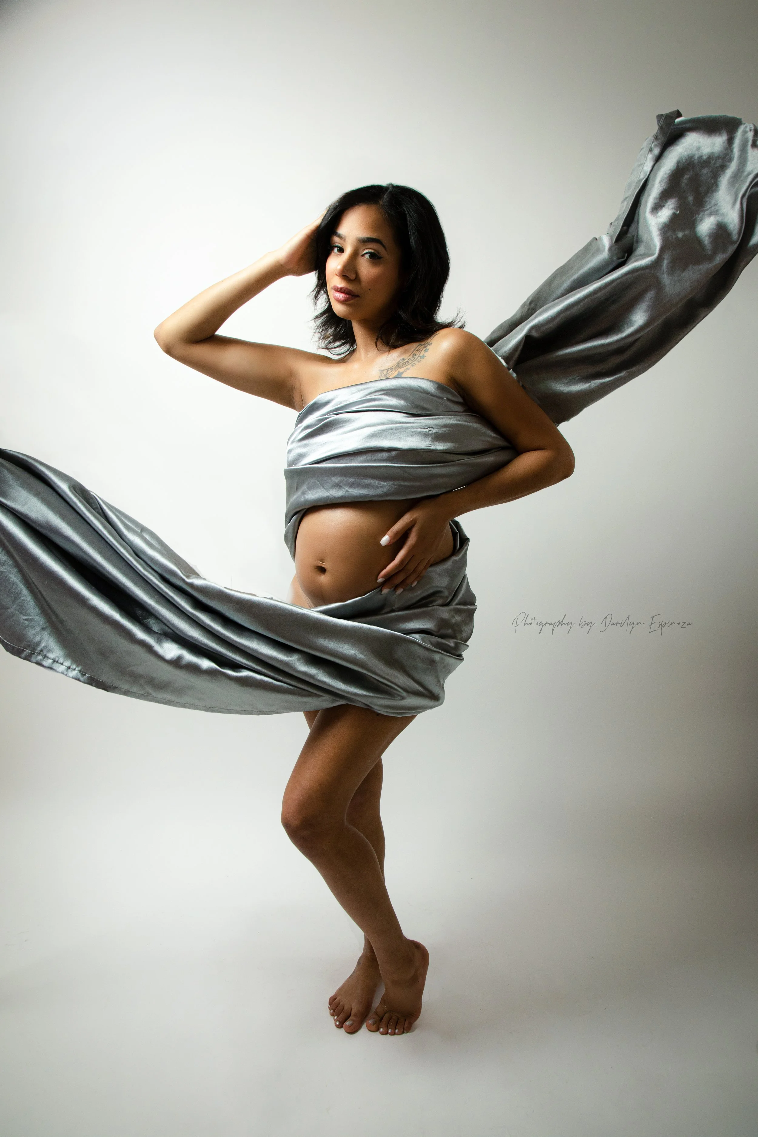 Pregnant woman wrapped in silver fabric against a plain background.