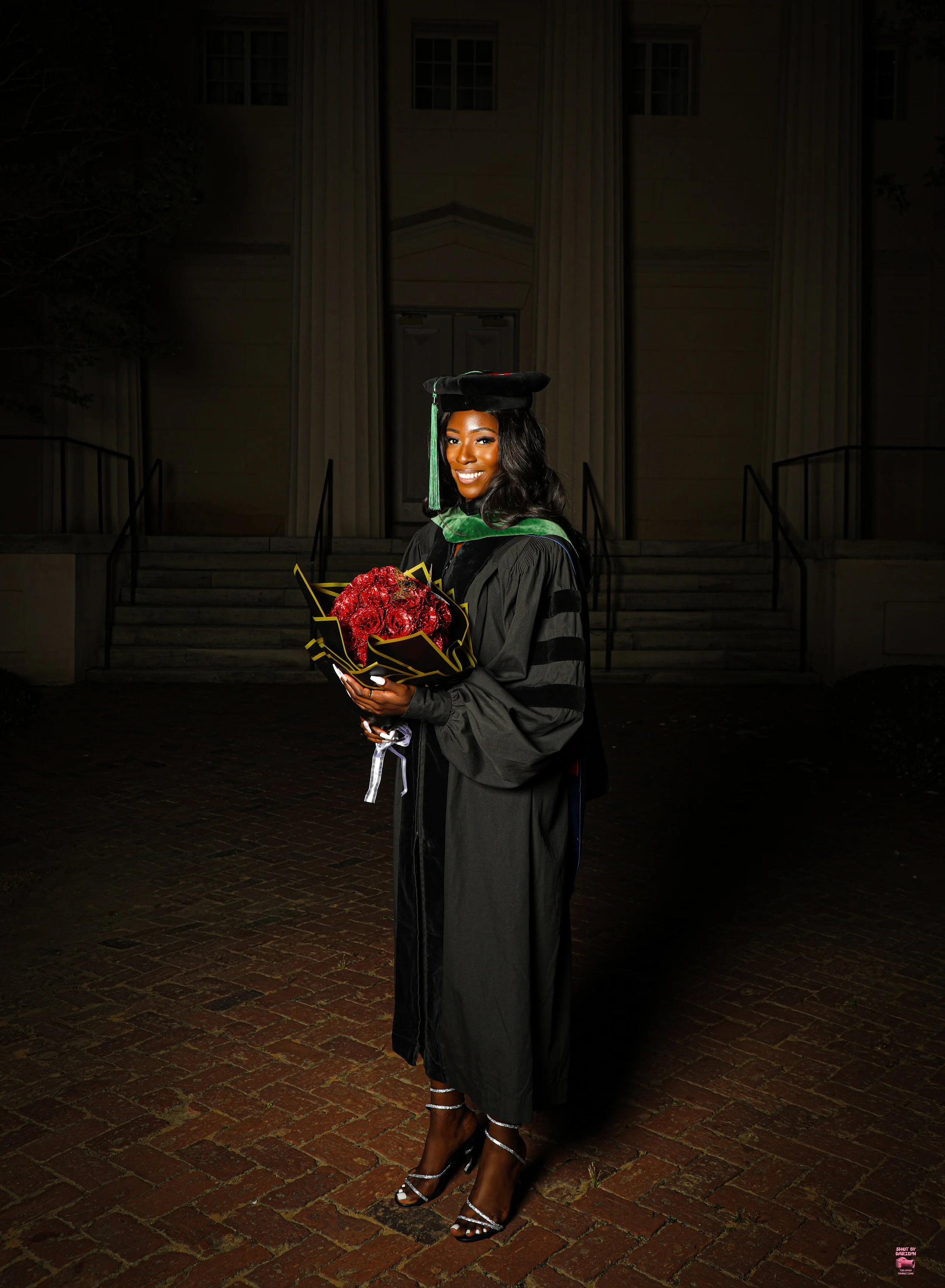 A woman in graduation attire, holding a bouquet of red flowers, standing outside at night in front of a building with steps.